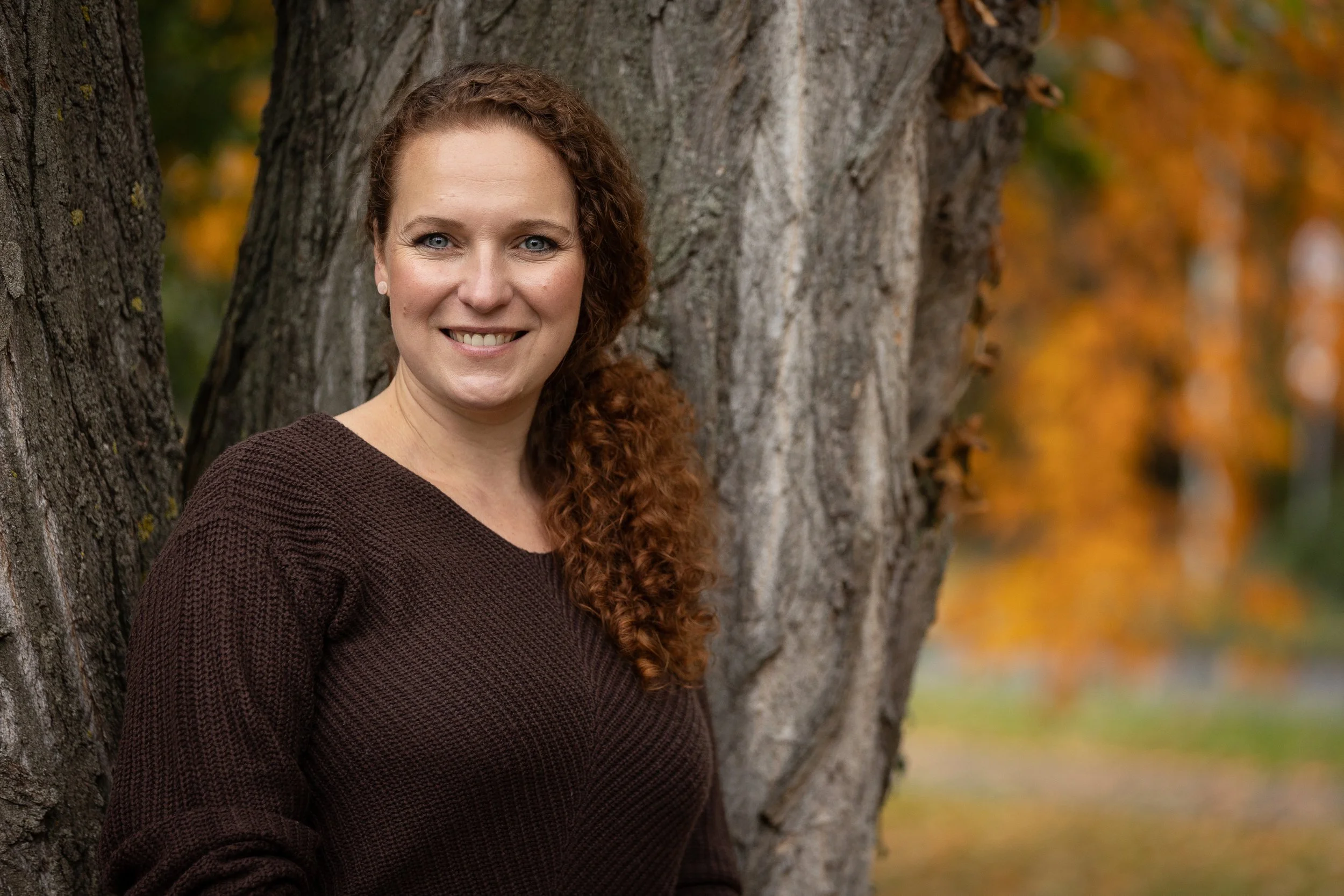 Eine lächelnde Frau mit roten, lockigen Haaren steht an einem Baum im Herbst mit orangefarbenen Blättern im Hintergrund.