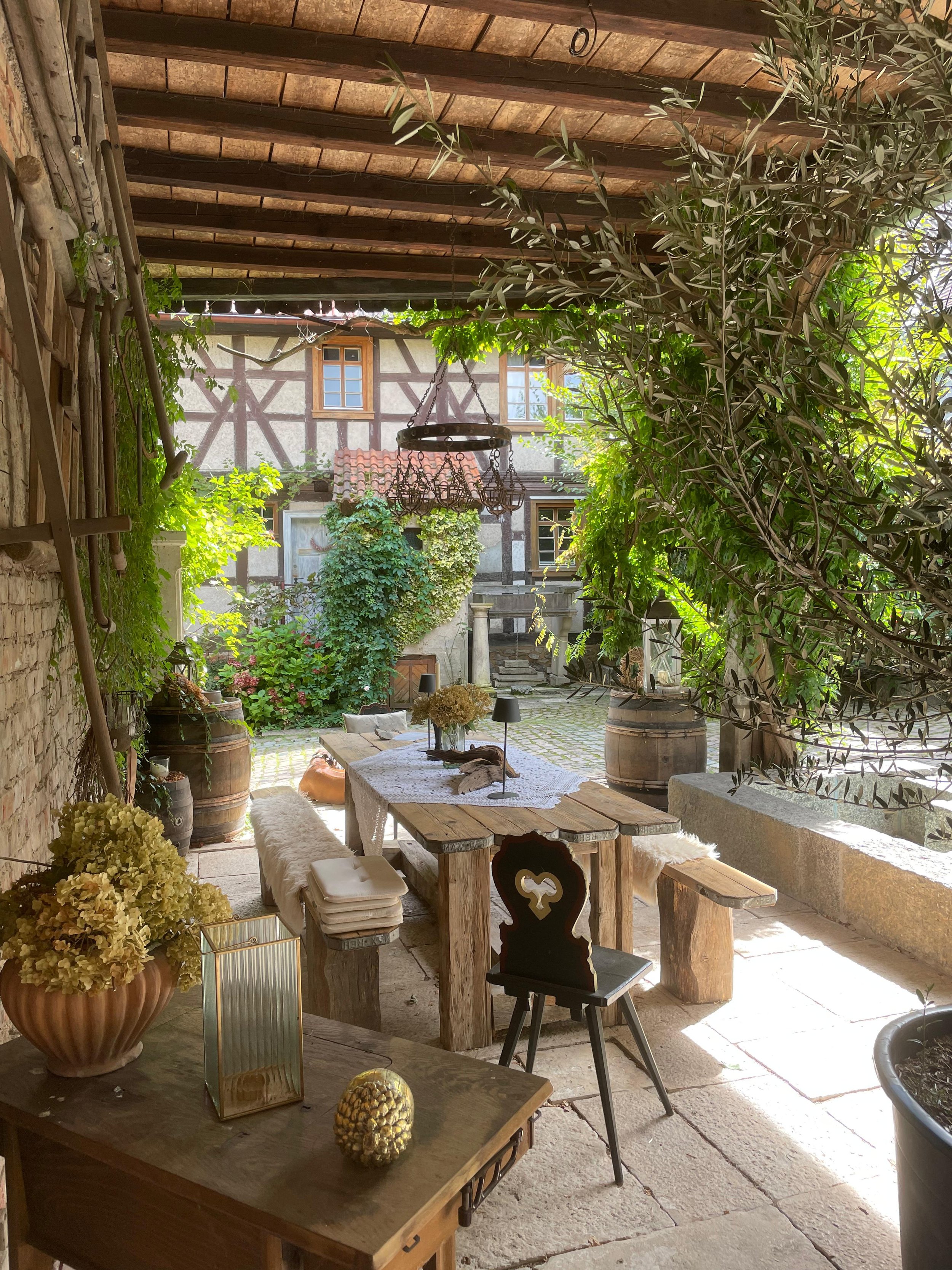 Ein dekorativer Hof mit rustikaler Einrichtung, Holztisch, Stühlen und Pflanzen, umgeben von einem Fachwerkhaus und einem geästen Garten, unter einem Holzdach.