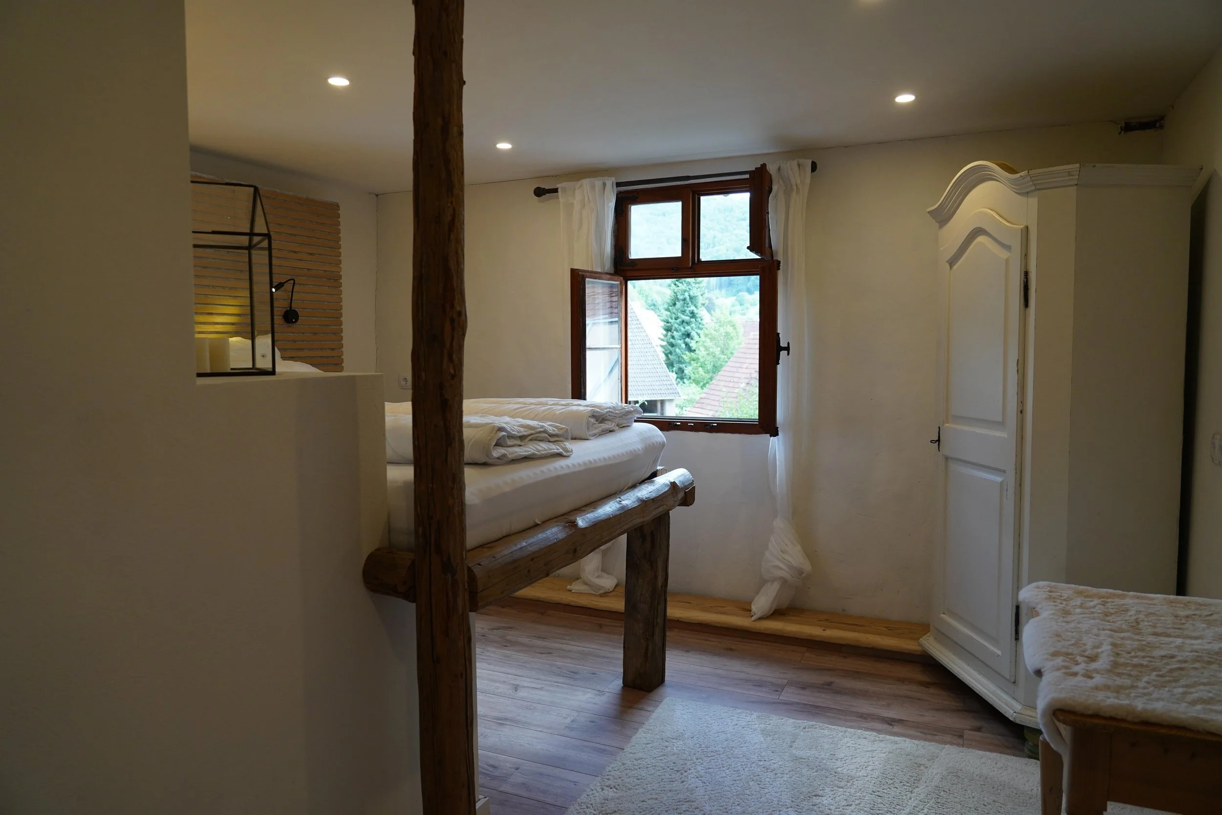 Ein gemütliches Schlafzimmer mit Holzfußboden, weißem Bett, einem offenen Fenster mit Blick auf grüne Bäscher und Hängelampen. Es gibt eine weiße Garderobe in der Ecke und eine kleine Ablage mit Handtüchern.