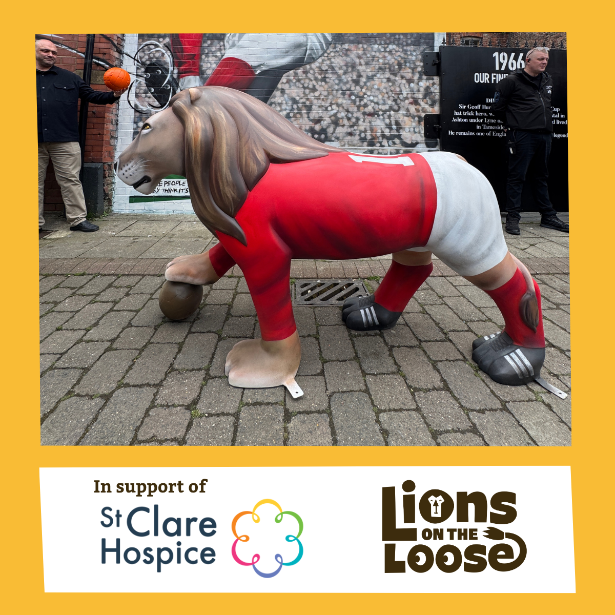 A lion sculpture dressed in a red sports jersey and white shorts, in a crouching position on a brick sidewalk, with a small brown ball under its paw, in front of a mural and two men standing nearby.