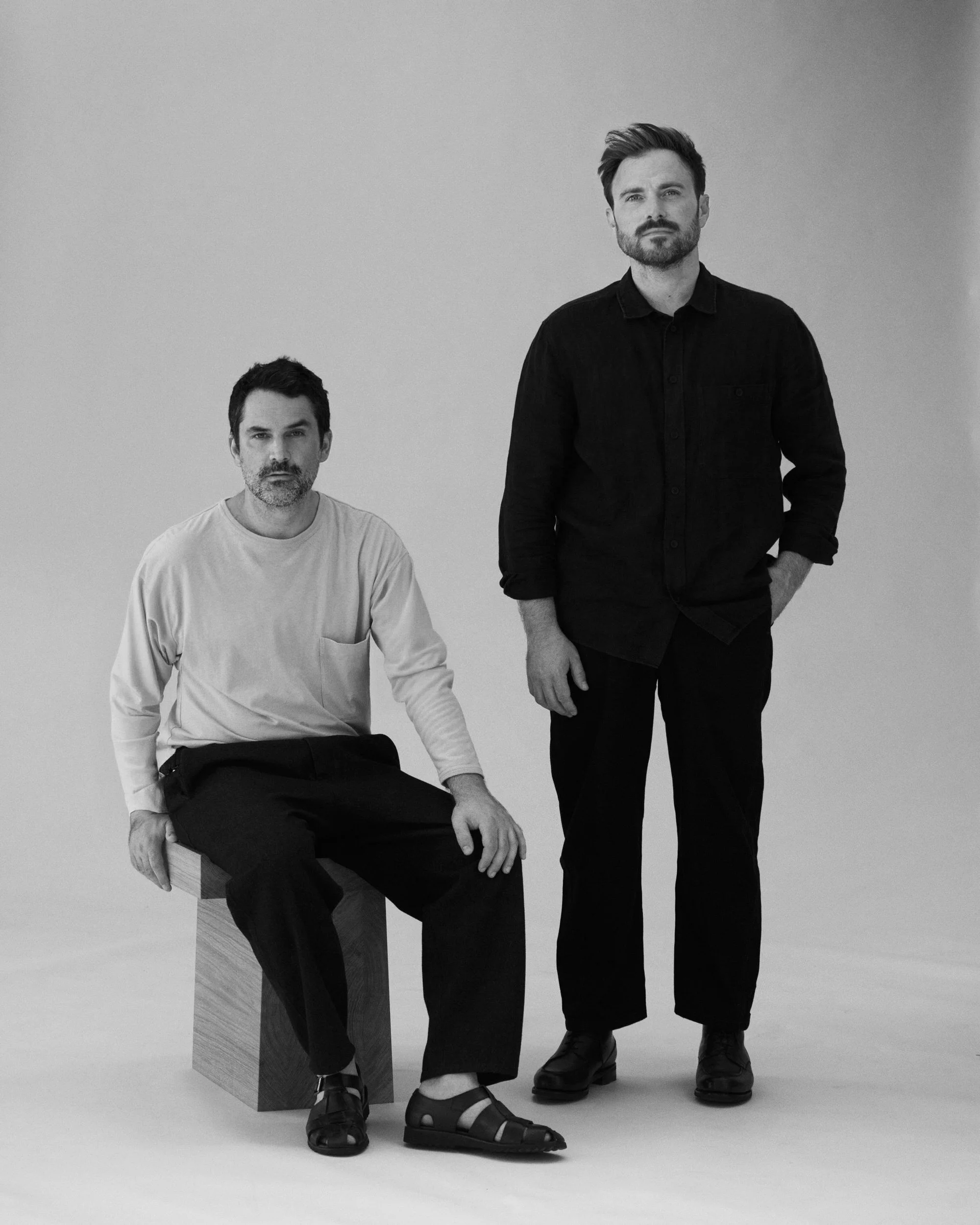 Photo studio en noir et blanc de Baptiste Rischmann et Guillaume Gibert. L'un assis sur un cube en bois, l'autre debout à côté.