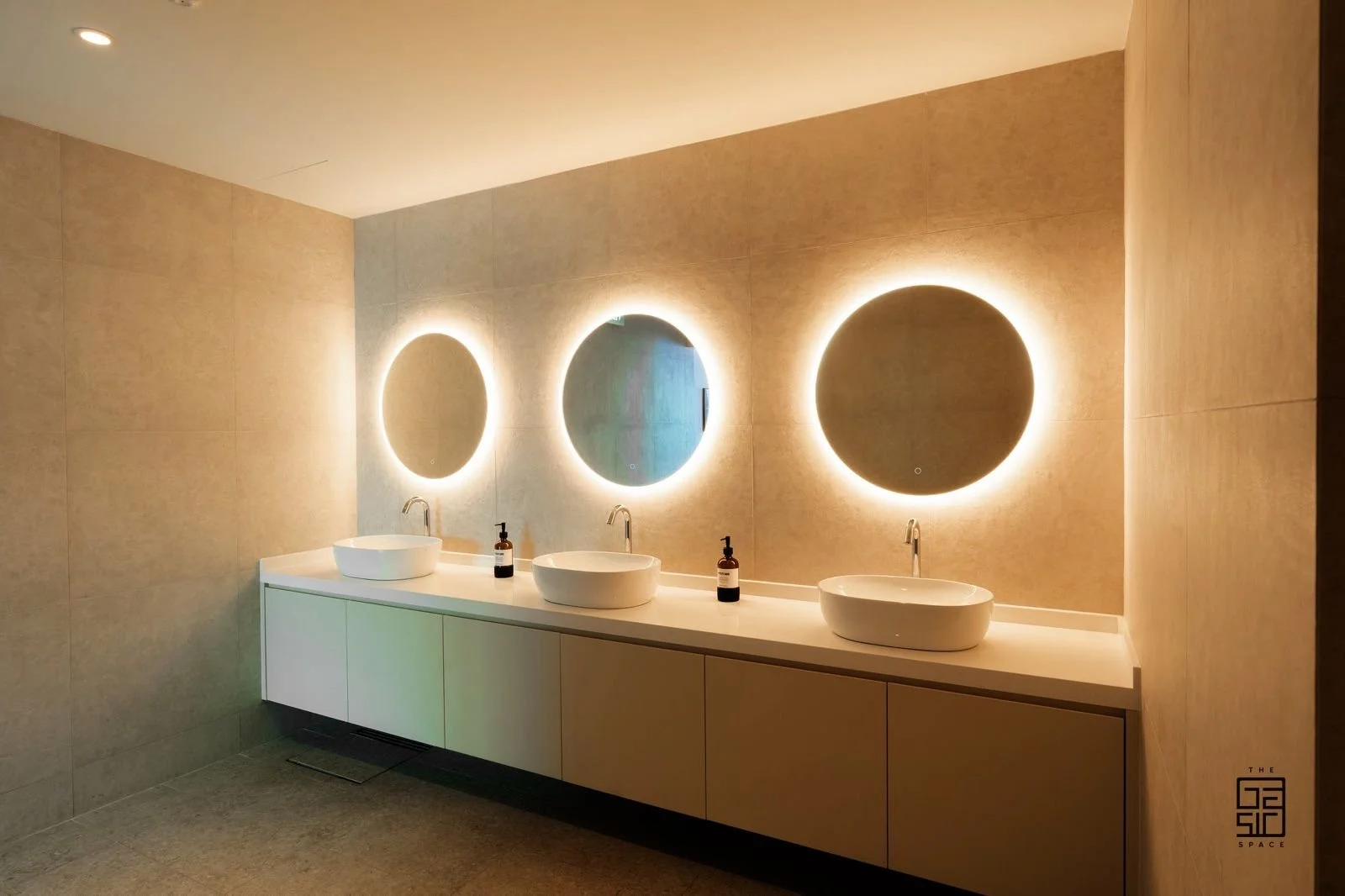 Modern bathroom with three white vessel sinks on a long white countertop, illuminated round mirrors with backlit lighting, beige tiled walls, and small soap dispensers between the sinks.