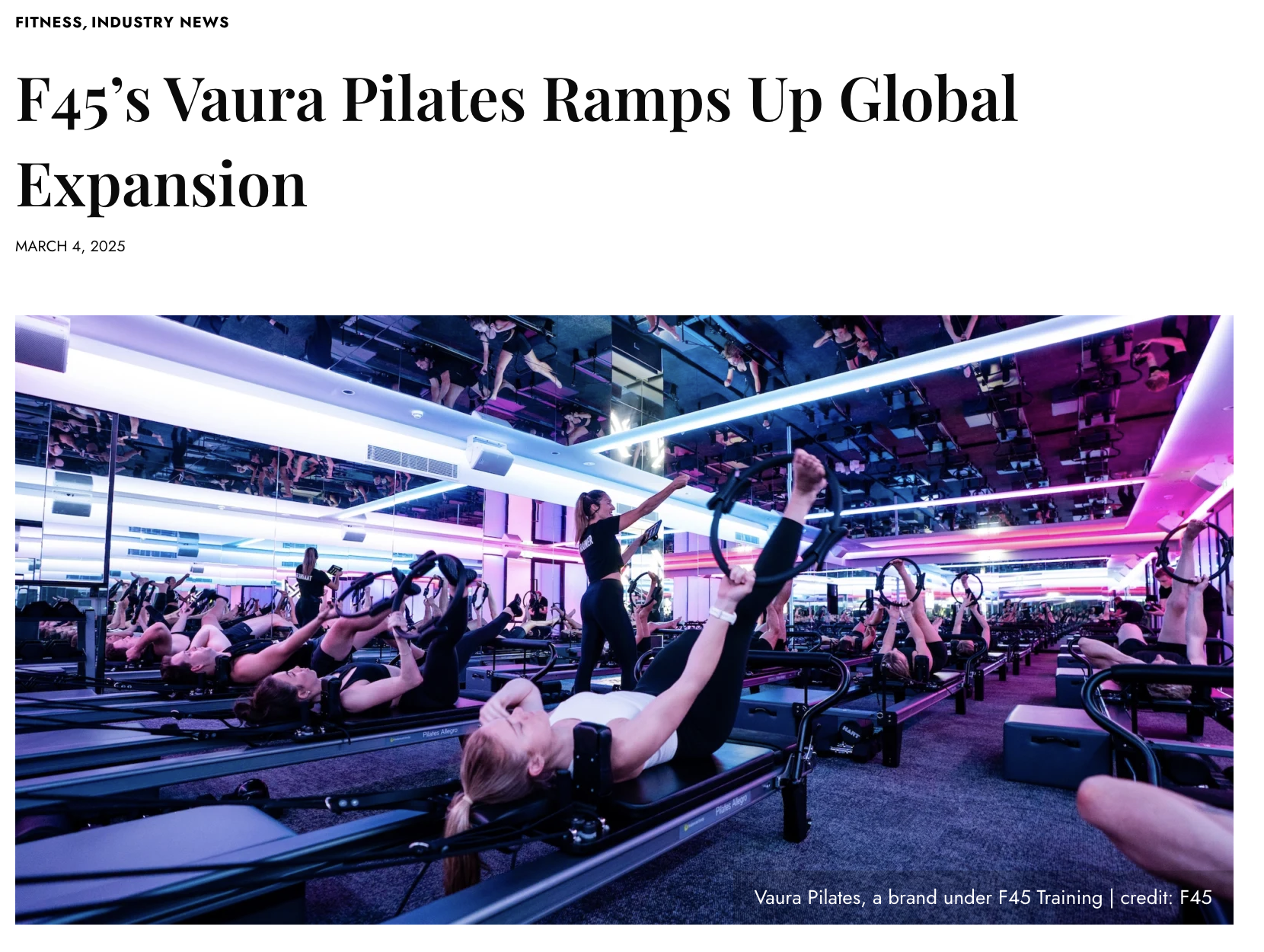 A Pilates class taking place in a brightly lit studio with blue and pink neon lighting, students lying on Pilates reformer machines and lifting their legs while an instructor guides them.