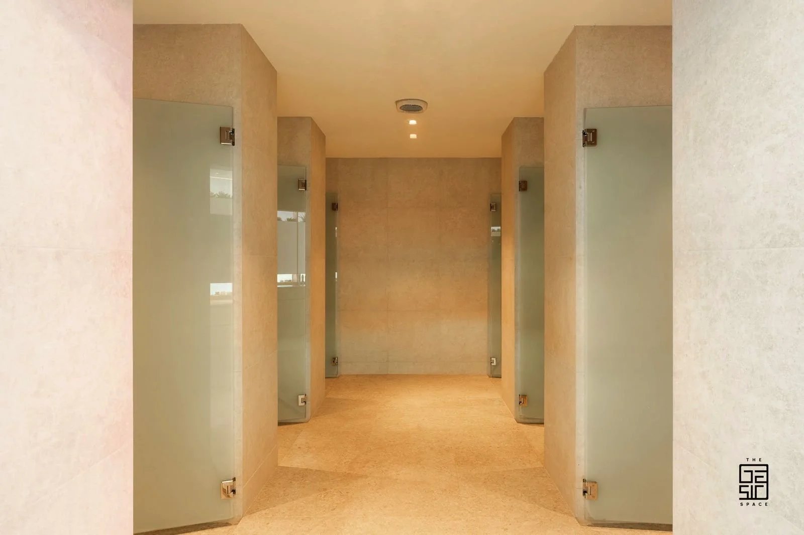 A public restroom with six stalls, each with frosted glass doors, beige tiled walls, and light-colored flooring.