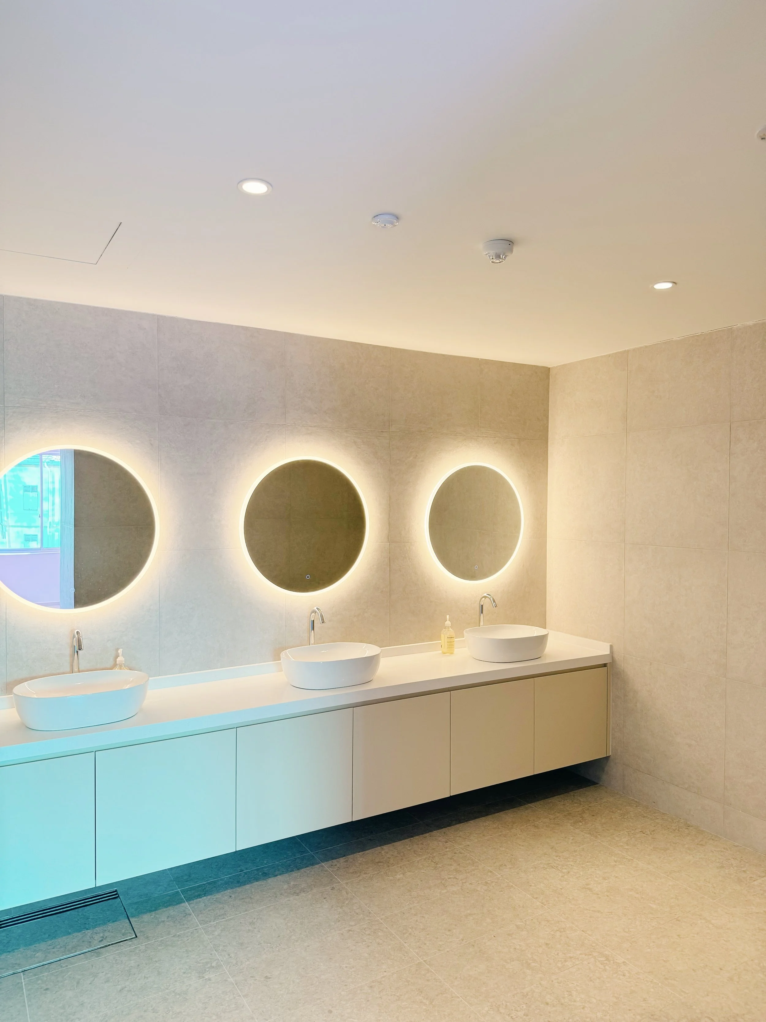 Modern public restroom with three illuminated circular mirrors above a white double vanity with two vessel sinks and a soap dispenser, beige tiled walls, and beige floor tiles.