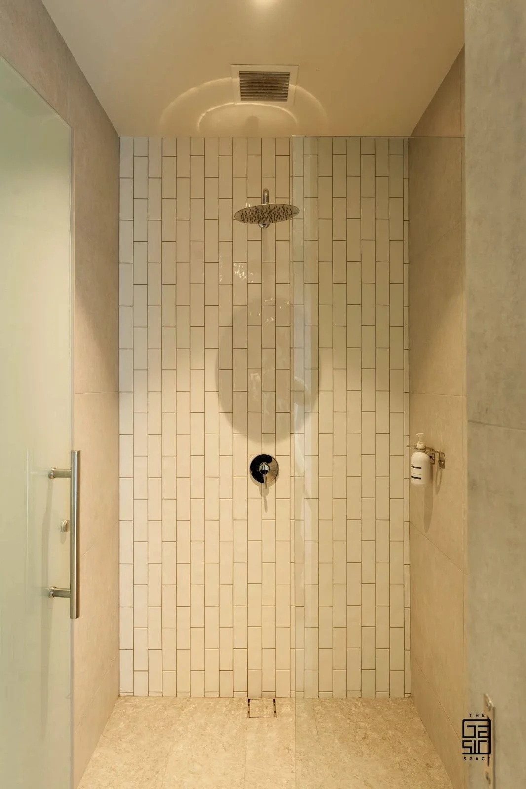 Empty shower with beige tiles, silver showerhead, black control, and soap dispenser on the wall.