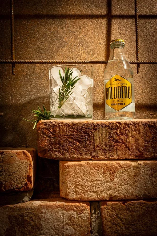 Produktfoto eines Longdrinks. Im Glas ist ein Rosmarinzweig, daneben steht eine Flasche Goldberg Tonic.