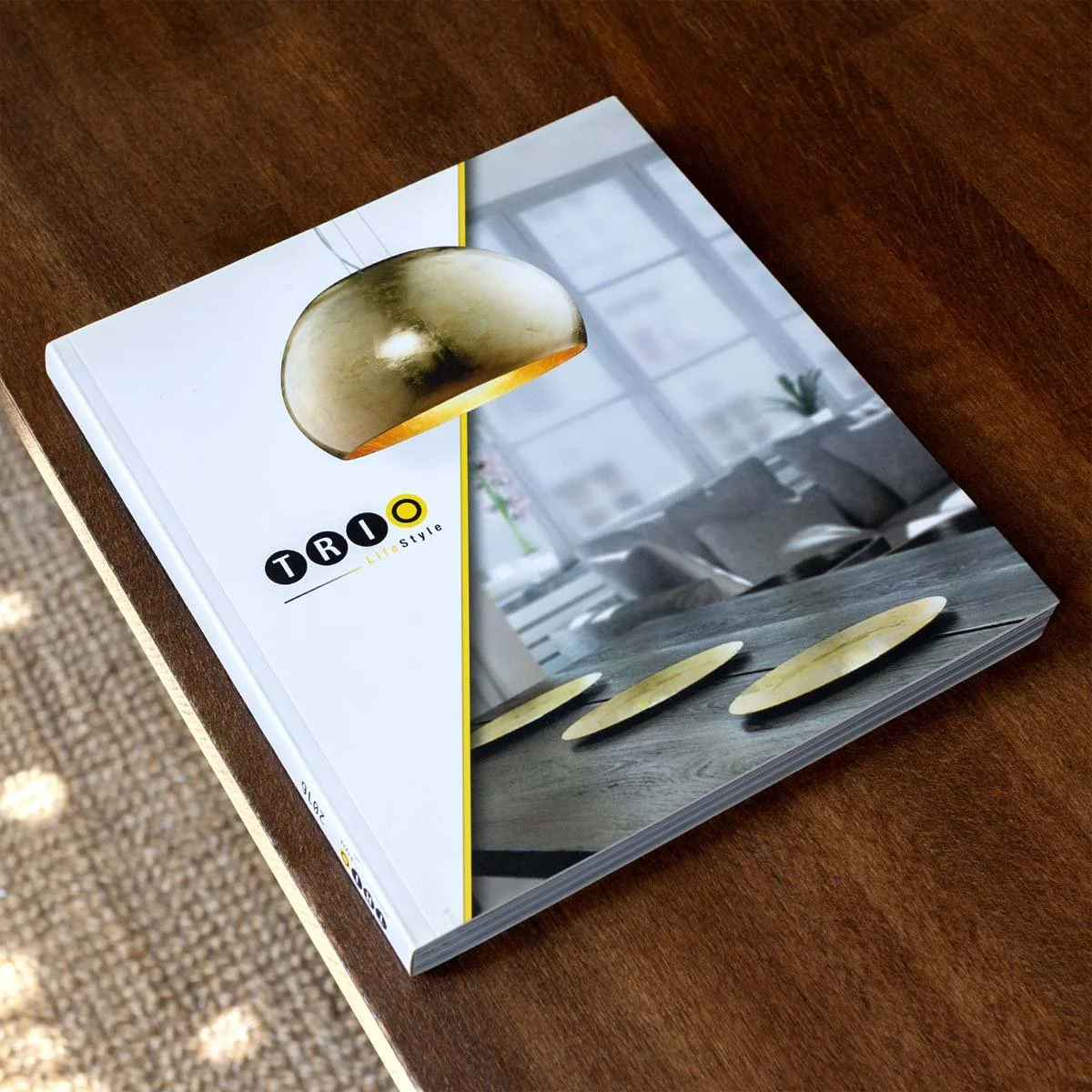 Katalog liegt geschlossen auf dem Tisch. Auf dem Titel ist das TRIO Logo zu sehen und eine goldene Pendelleuchte.