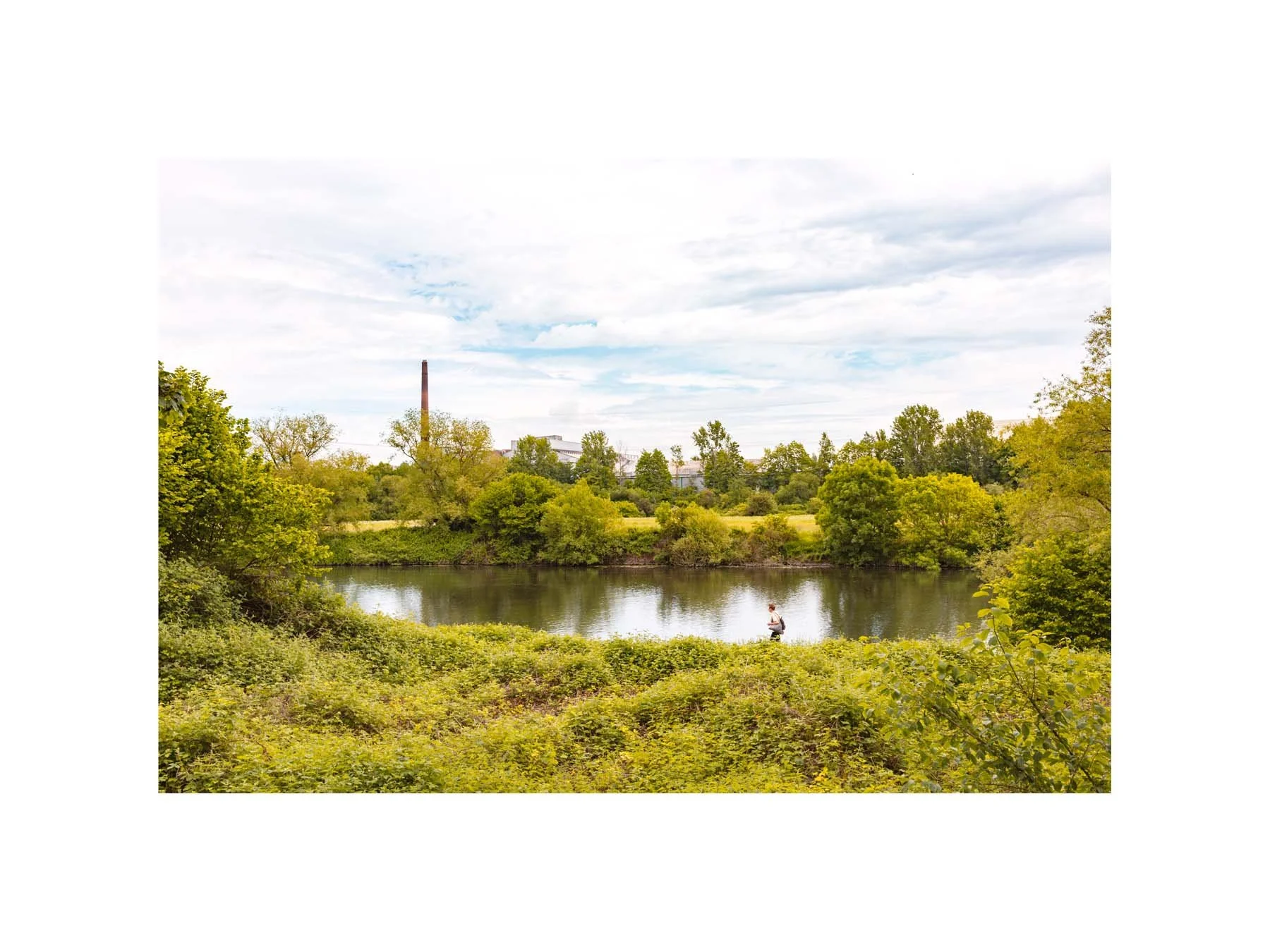 Foto von der Ruhr. Das Foto trägt den Titel "Gefühl von Ruhr" und man sieht die Ruhr, eingeschlossen von grünen Sträuchern. Im Vordergrund läuft ein Mensch von rechts nach links und im Hintergrund sehen wir einen Schornstein einer alten Fabrik.