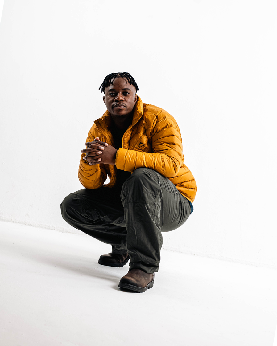 A man crouching with hands clasped, wearing a yellow puffer jacket, black pants, and brown boots against a white background.