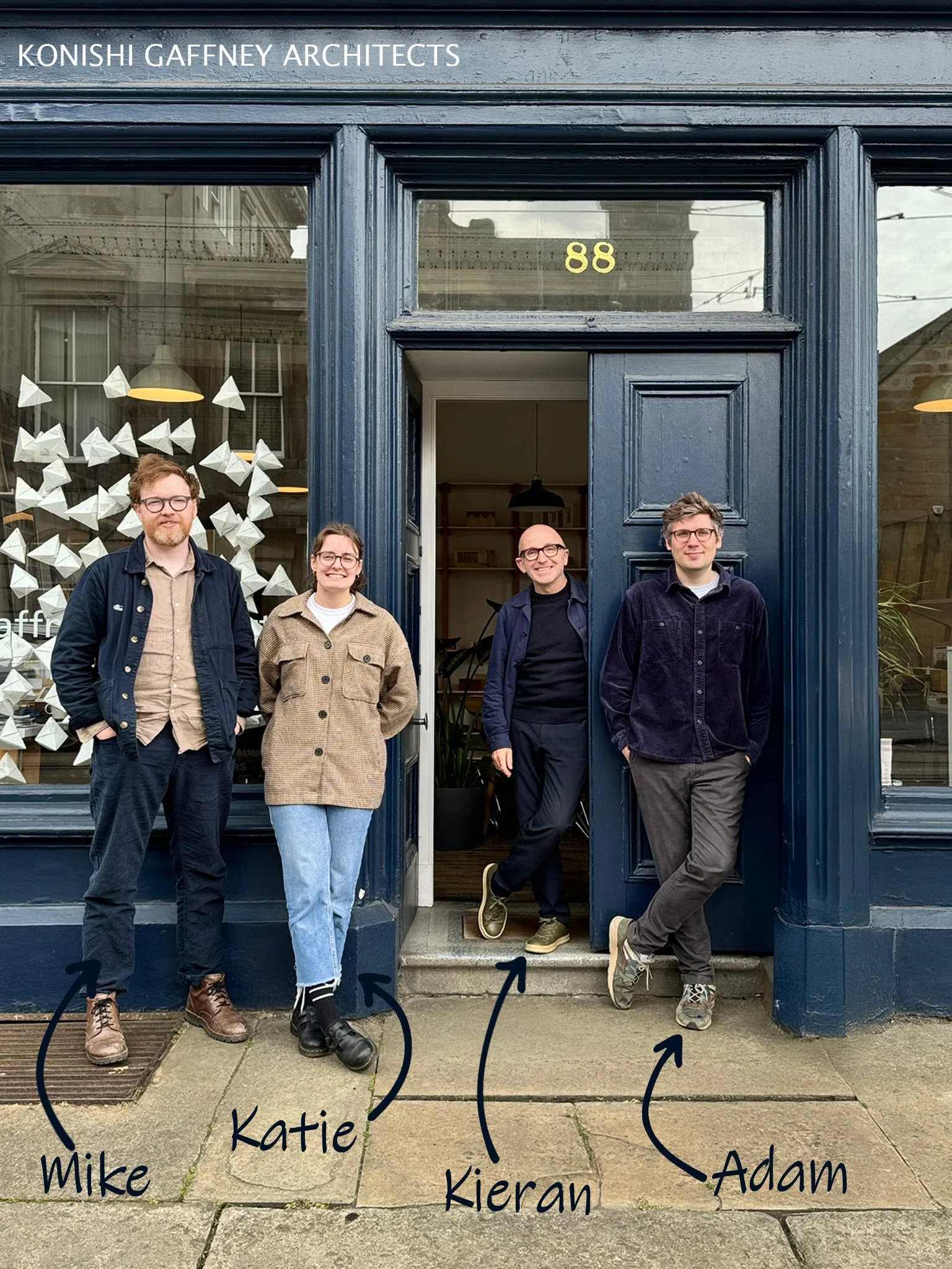 Four people standing outside a dark blue building with the address number 88 above the door, smiling. The building has large windows displaying art and lighting inside. The people from left to right are labeled as Mike, Katie, Kieran, and Adam.