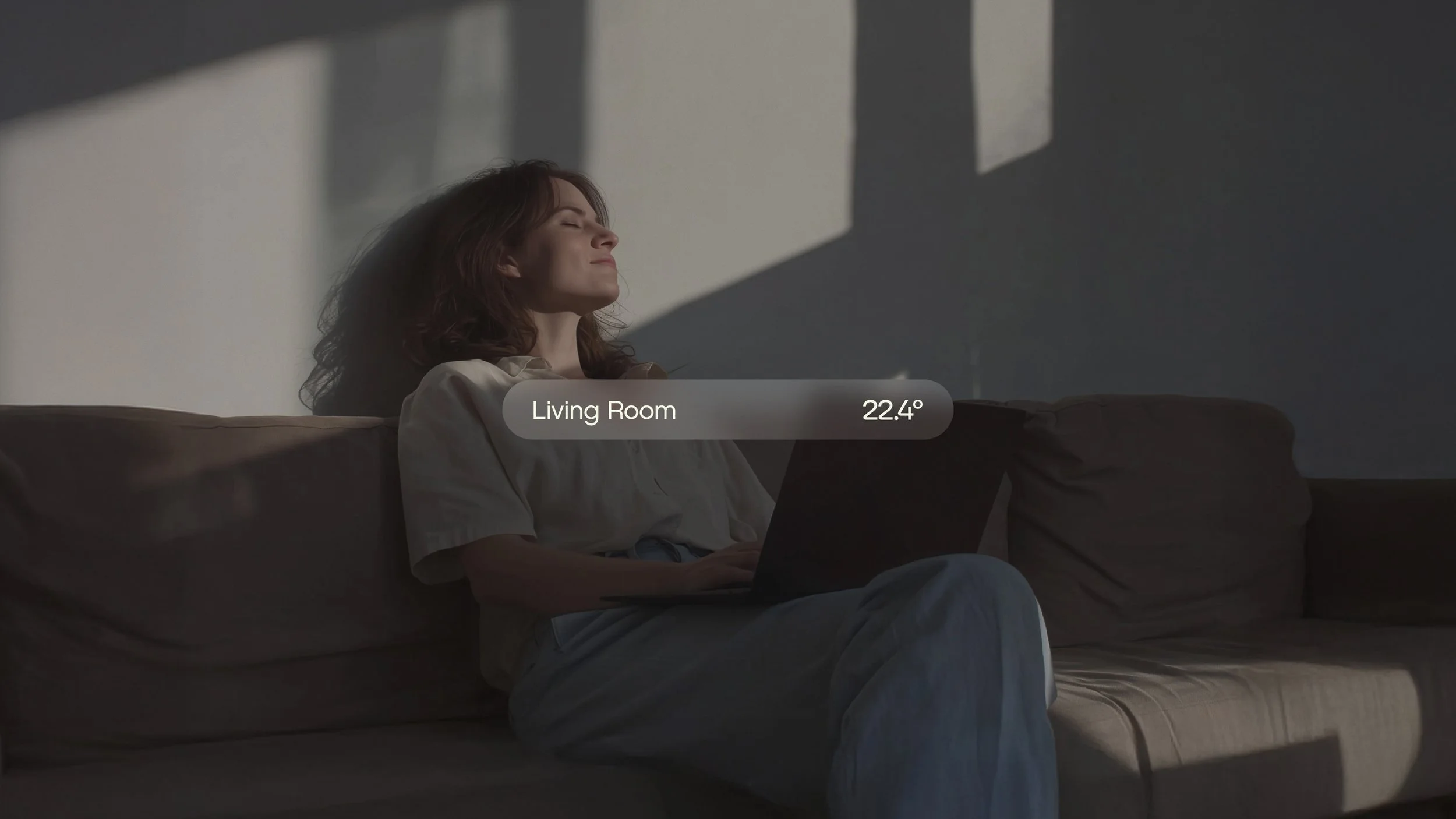 Woman sitting on a beige couch with a laptop, eyes closed, sunlight casting shadows on the wall, temperature reading 'Living Room 22.4°'
