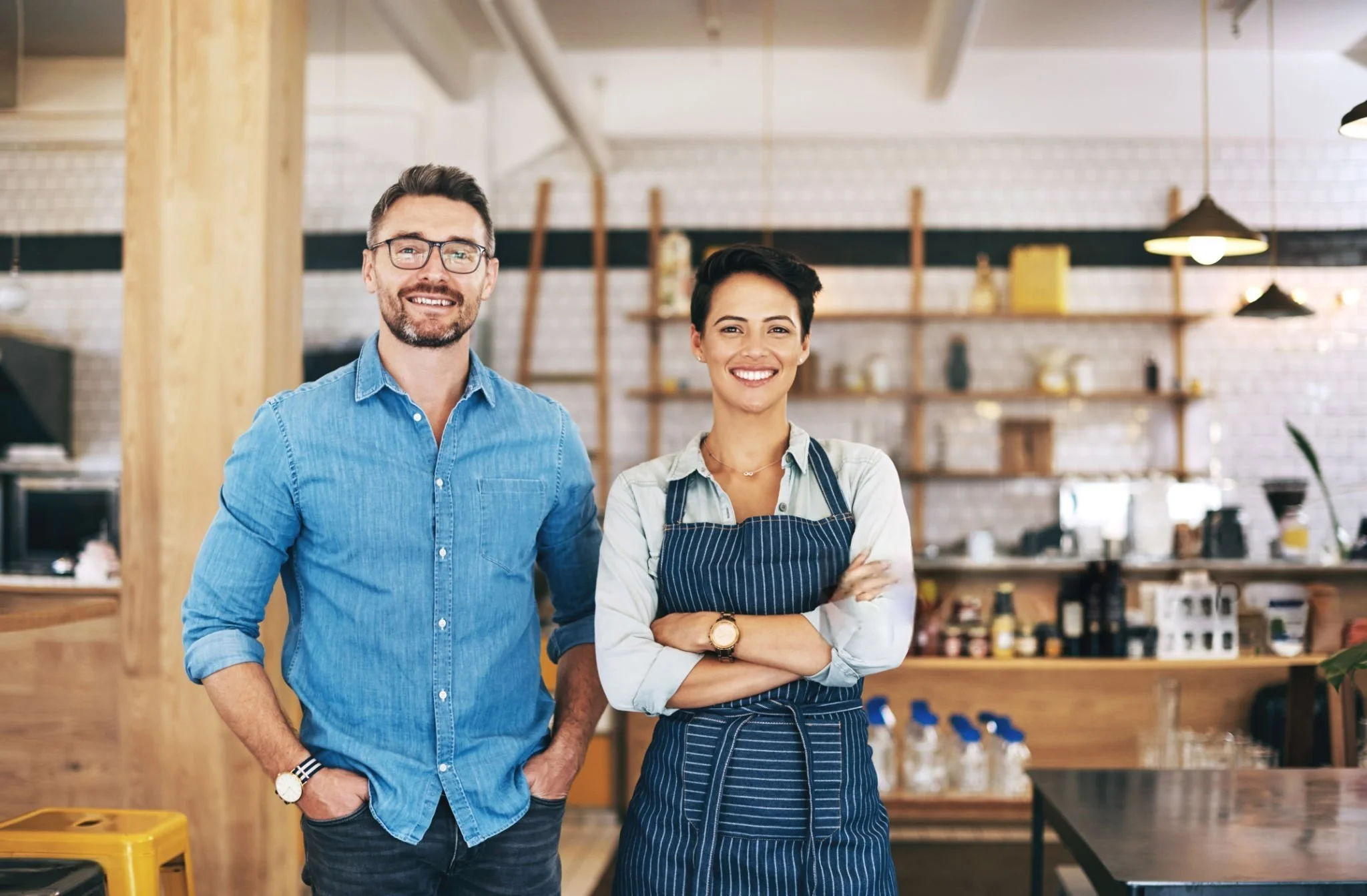 A man and woman smiling in a cafe or restaurant setting, with the woman wearing an apron.