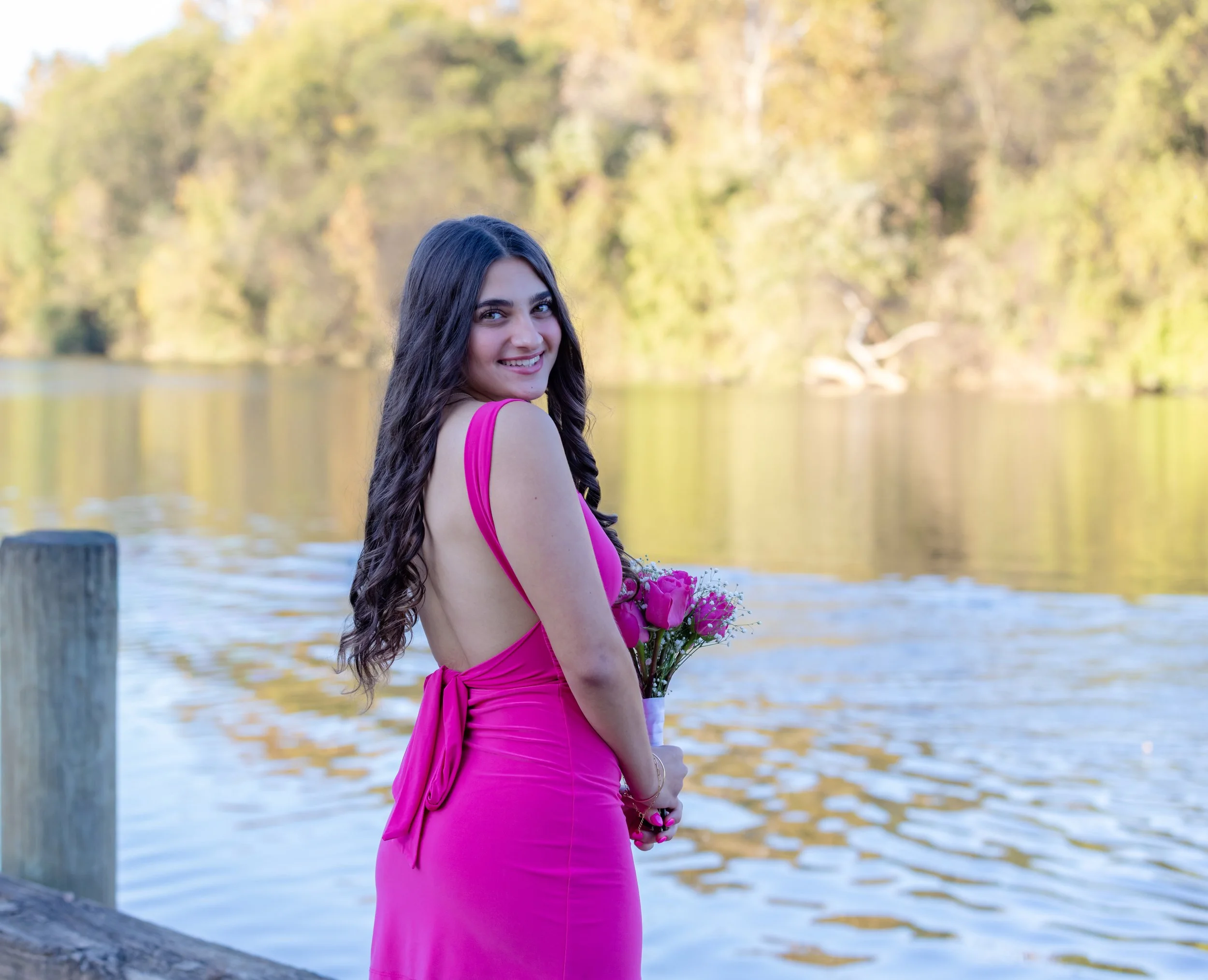Young woman in a pink dress holding a bouquet of flowers by a lake with trees in the background.