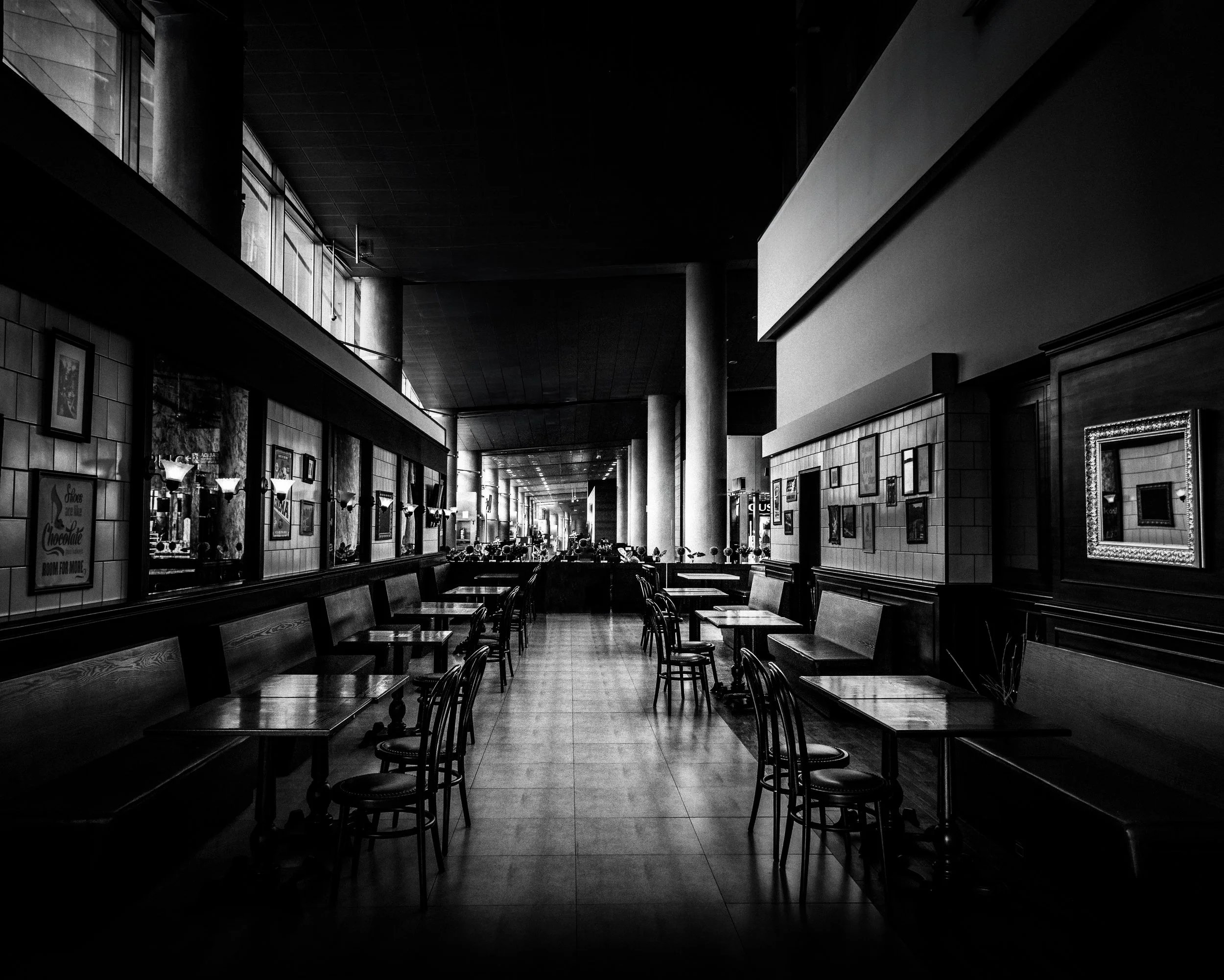Empty restaurant with chairs and tables, framed pictures on the walls, and light coming from windows.