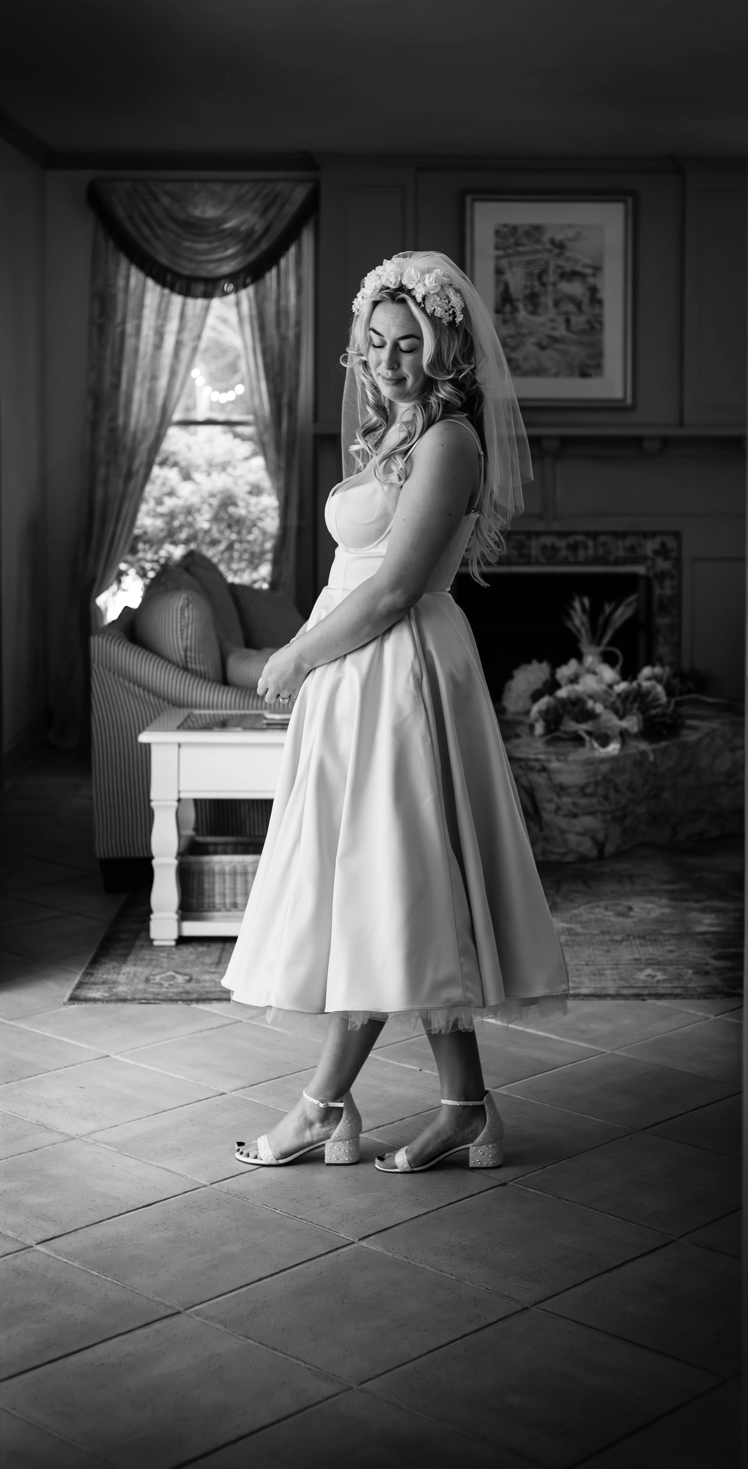 Black and white photo of a bride in a room with a window, dressed in a wedding gown and heels, standing with eyes closed and a gentle smile.