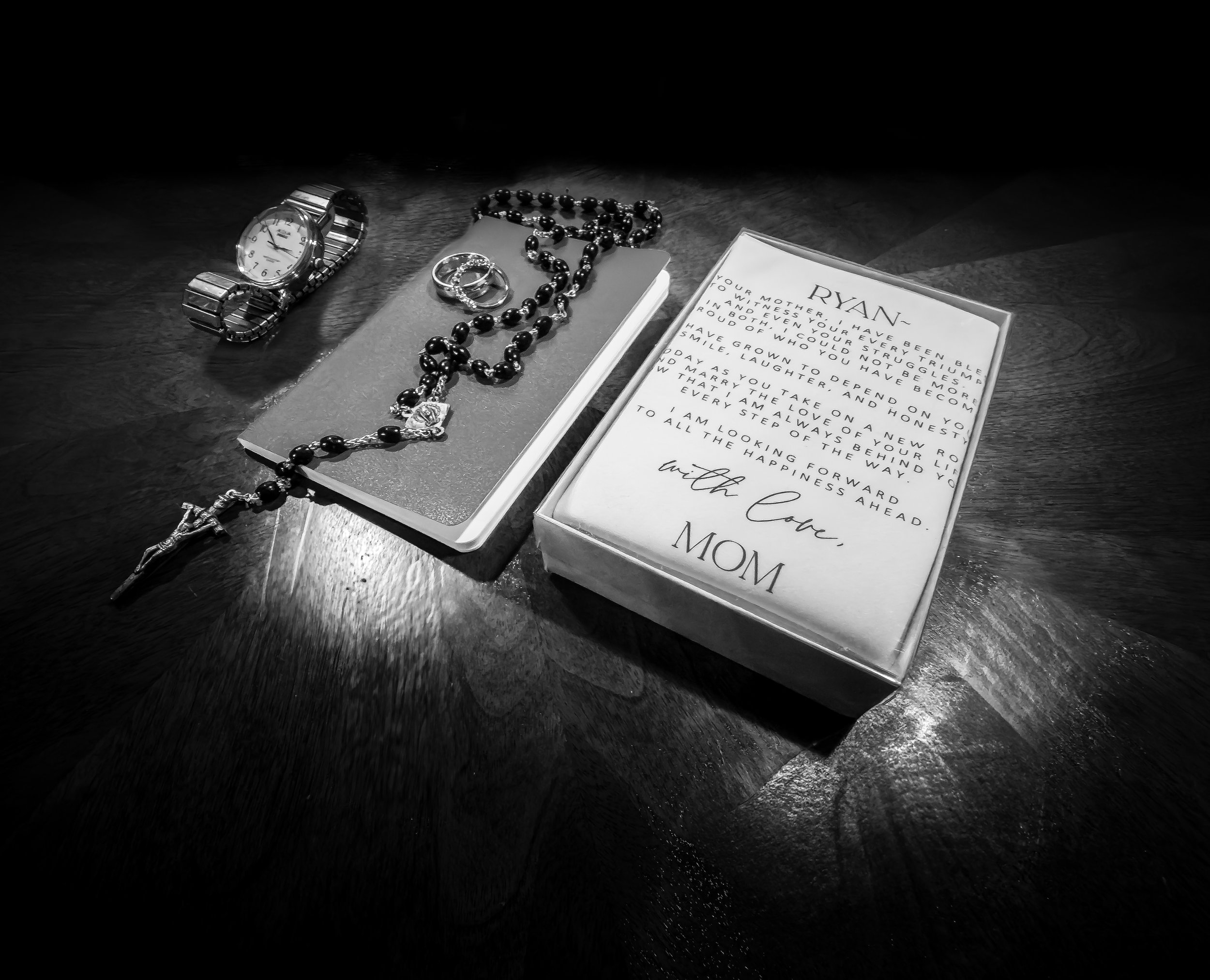 A wristwatch, rosary beads, and rings on a small book, with a card containing a heartfelt message from a mom to her son, on a wooden surface.