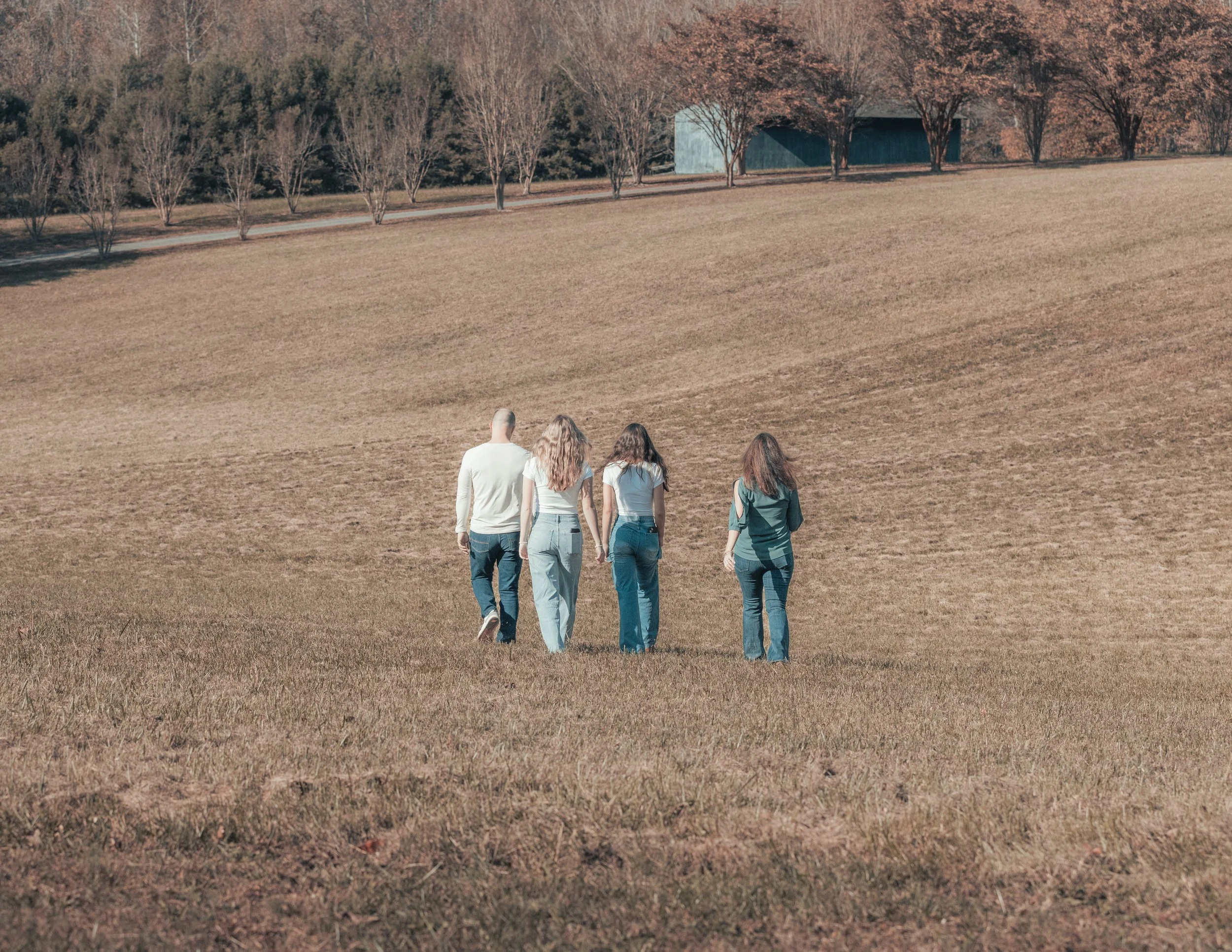 Four people walking away across a grassy open field, with trees and a small building in background.