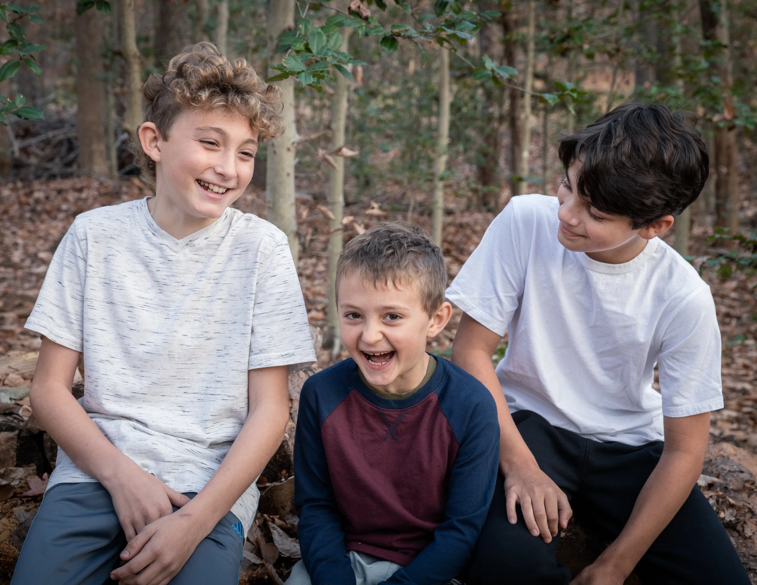 Three boys sitting outdoors in a wooded area, laughing and smiling.