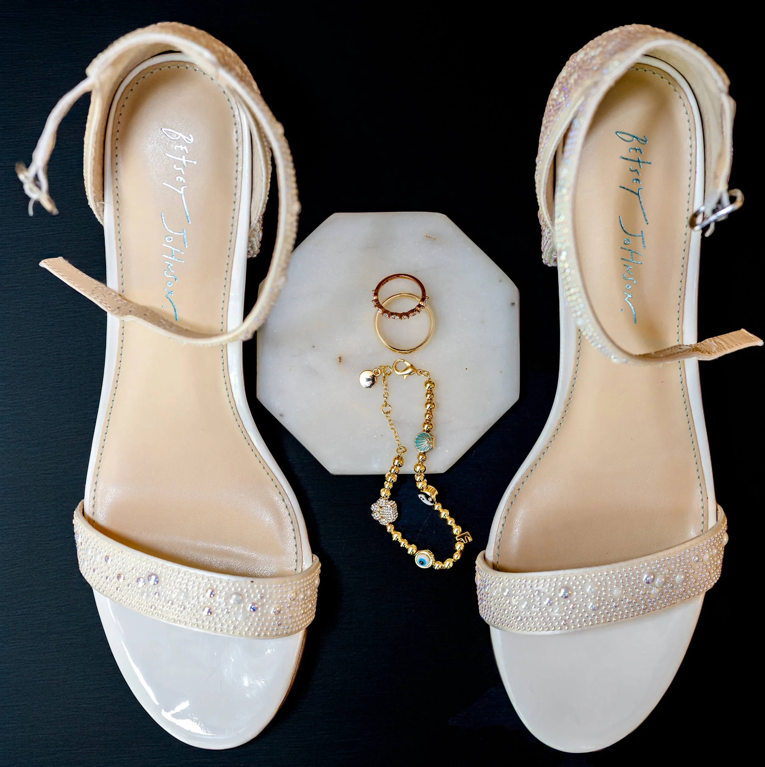 Pair of white high-heeled sandals with pearl and rhinestone embellishments, gold rings, a bracelet with charms, and a marble coaster underneath, on a black surface.