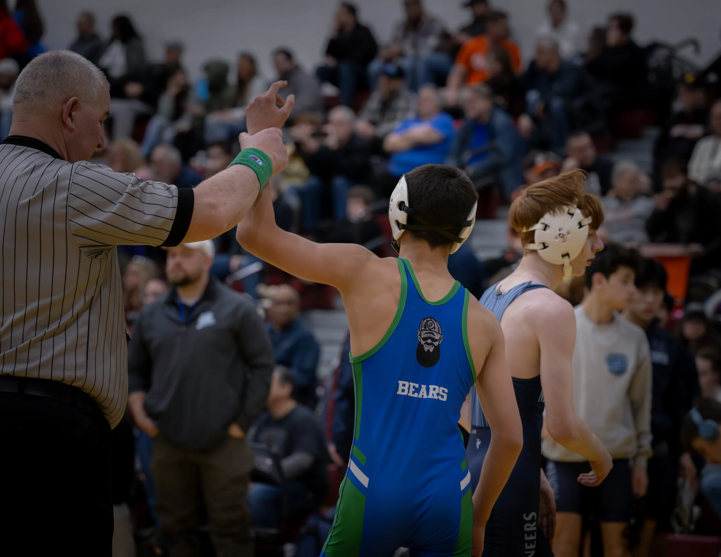 Two young wrestlers in singlets and protective headgear receive a sportsmanship handshake from a referee during a wrestling match in a gym filled with spectators.