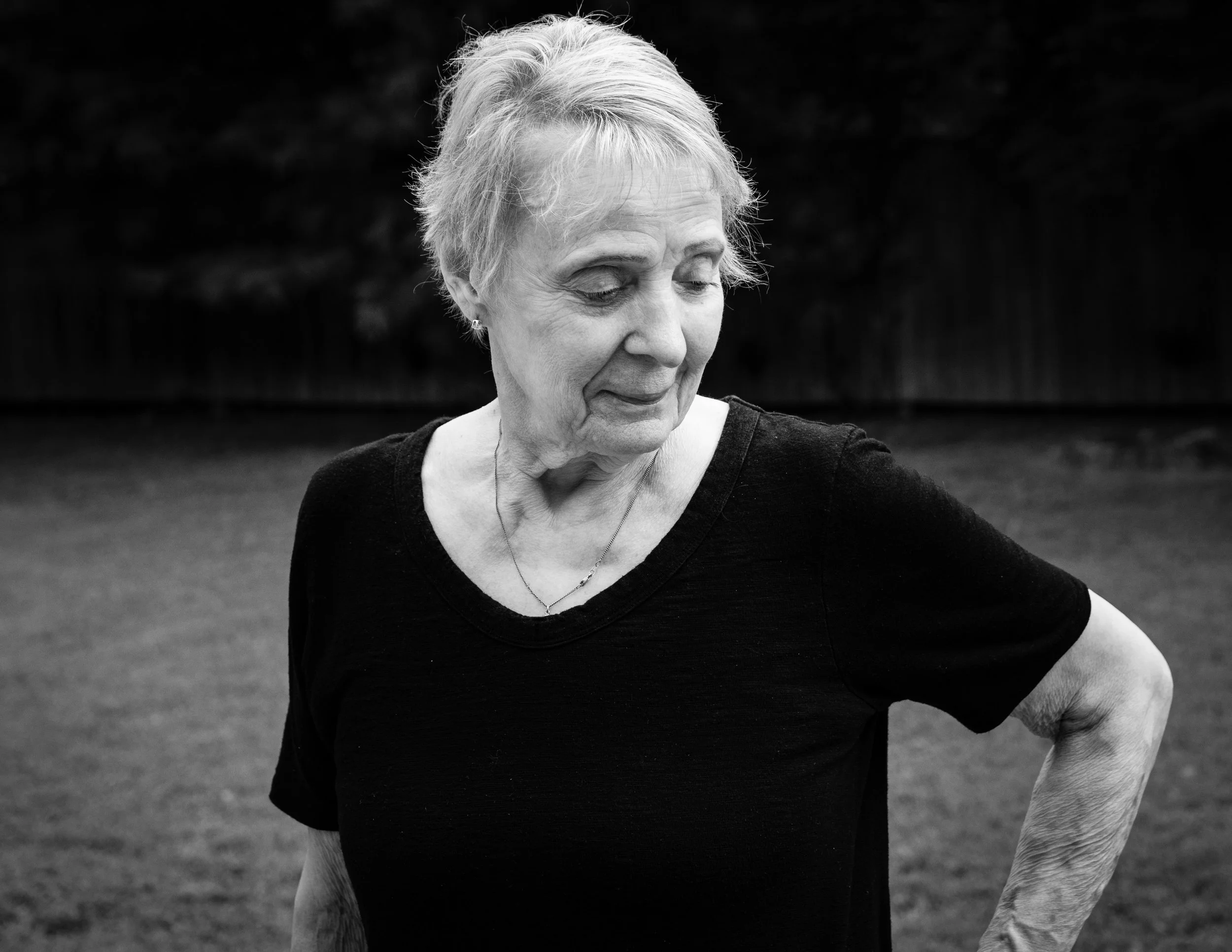 A black and white photo of an elderly woman with short hair, wearing a black shirt, standing outdoors on a grassy field with a fence and trees in the background. She has her head tilted downward with her eyes closed and her right hand on her hip.