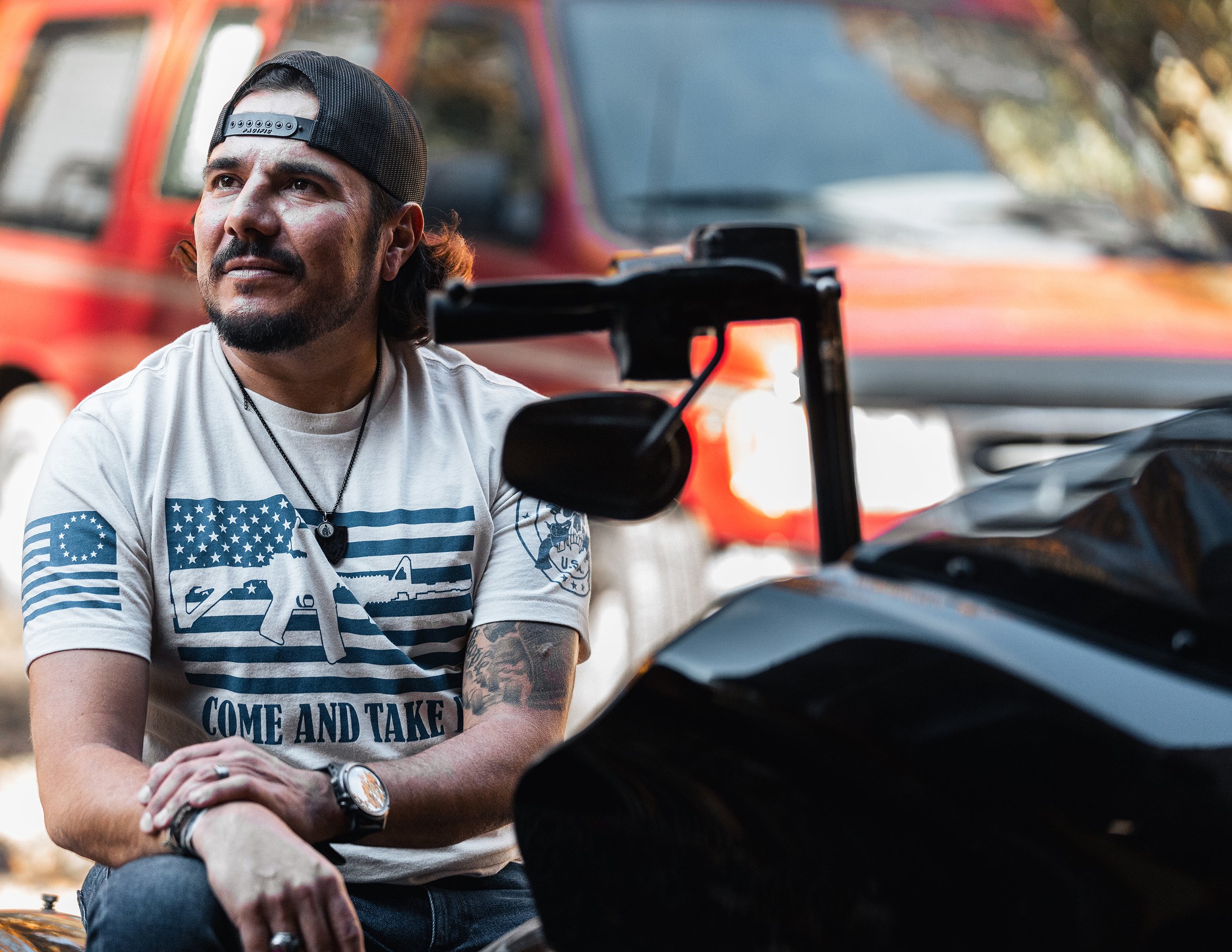 A man with a beard wearing a black cap backwards, a white t-shirt with American flag-themed graphics, and a necklace, is sitting outdoors near a motorcycle with a blurred red vehicle in the background.
