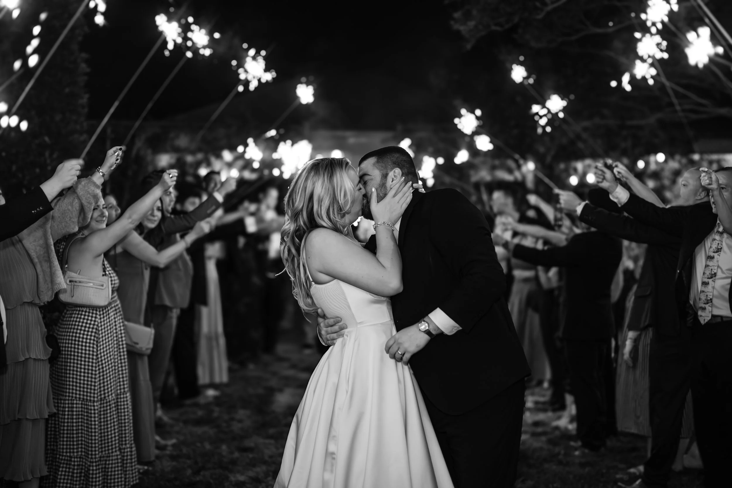 Black and white photo of a bride and groom kissing at their wedding reception under string lights, surrounded by guests holding sparklers.