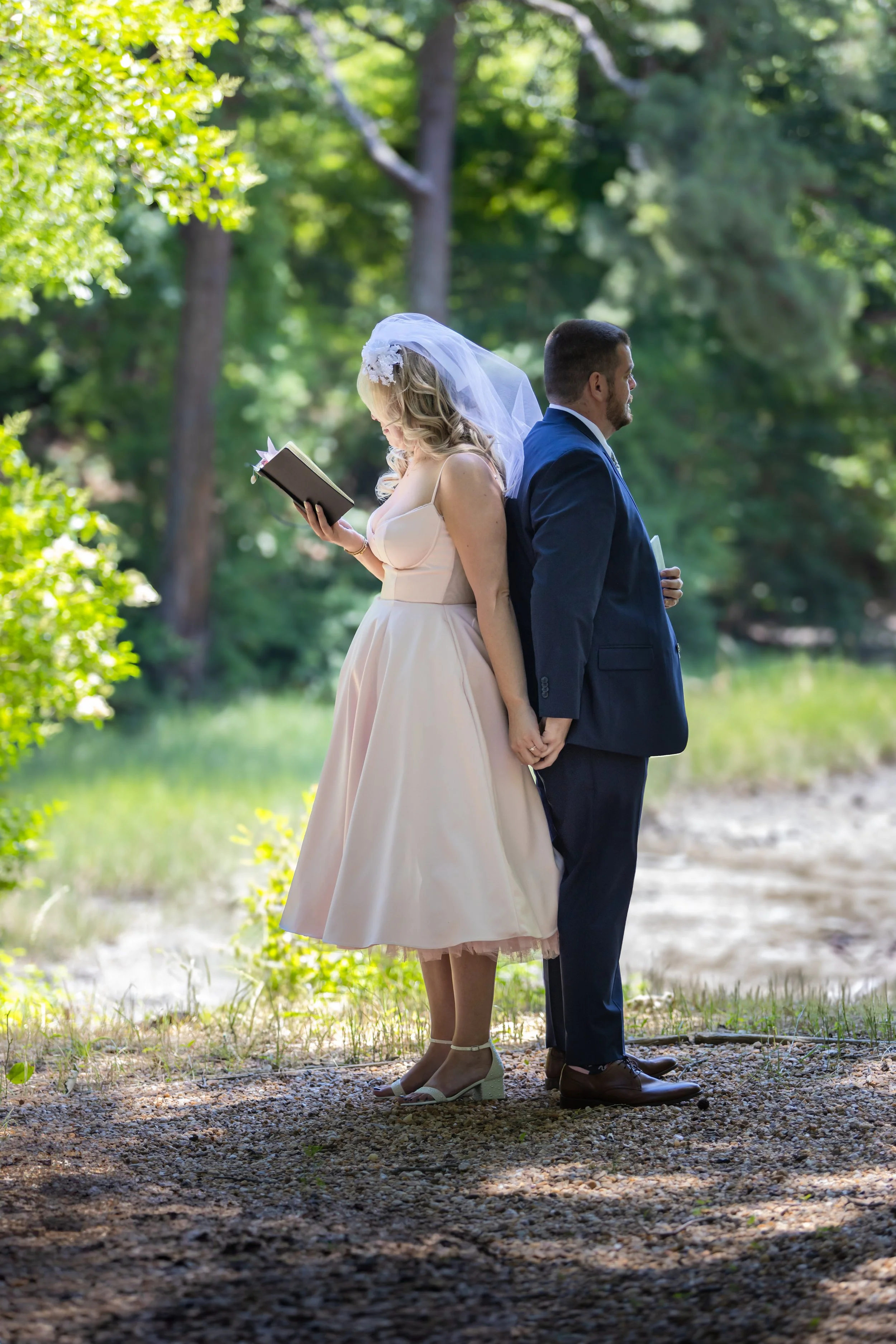 Bride and groom standing back to back outdoors, holding hands, with bride reading a book, surrounded by green trees and sunlight.