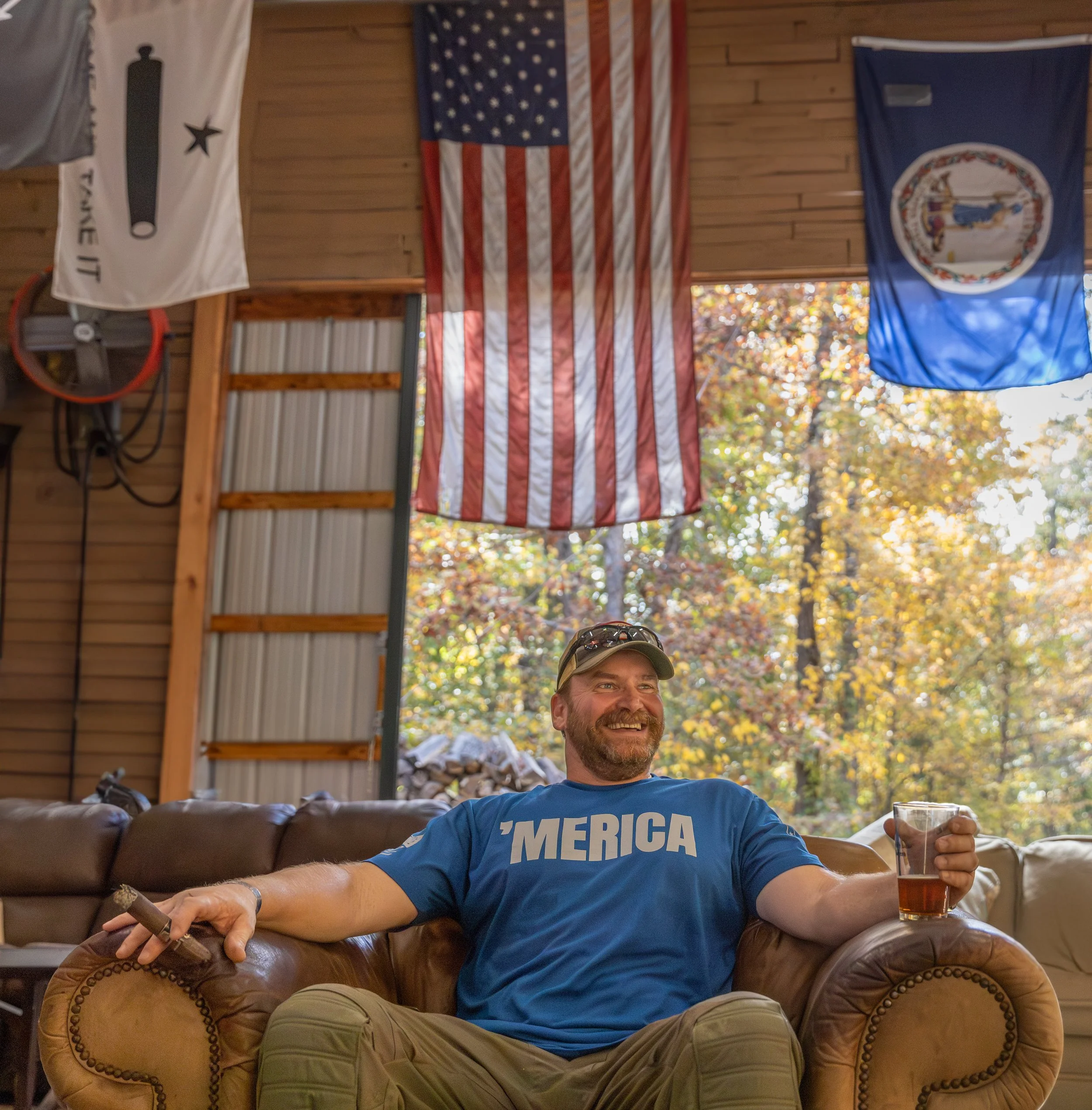A man with a beard sitting on a brown leather couch, smiling and holding a cigar in one hand and a glass of dark beverage in the other. Behind him, an American flag and another flag hang from the ceiling, with autumn trees visible outside.