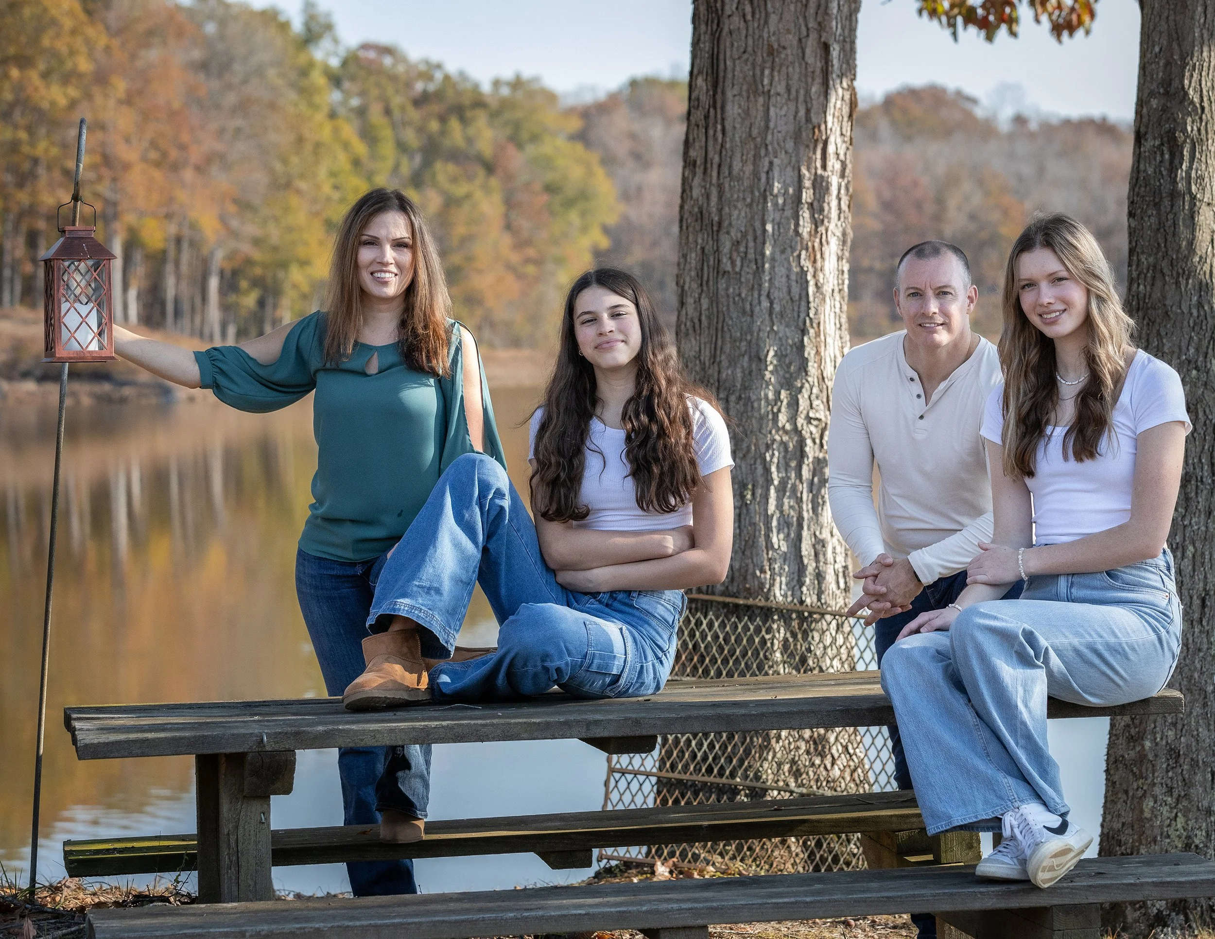 A family of four posing outdoors by a lake with autumn trees in the background. They are sitting and standing on a wooden dock, dressed casually, with two women and one man smiling at the camera.