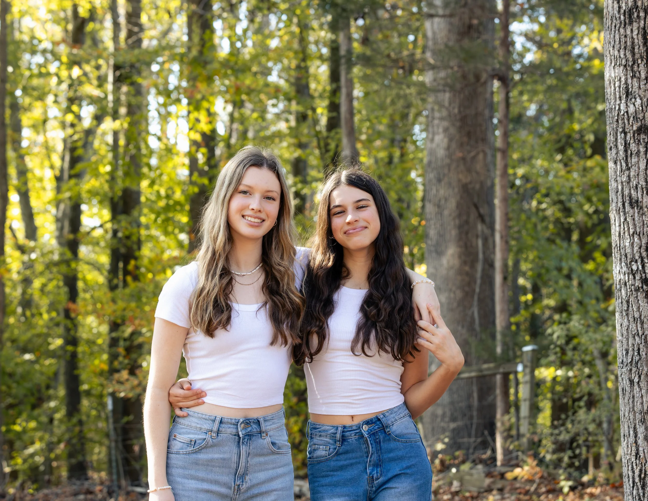 Two young women with long hair standing outdoors in a wooded area, smiling and arm-in-arm.