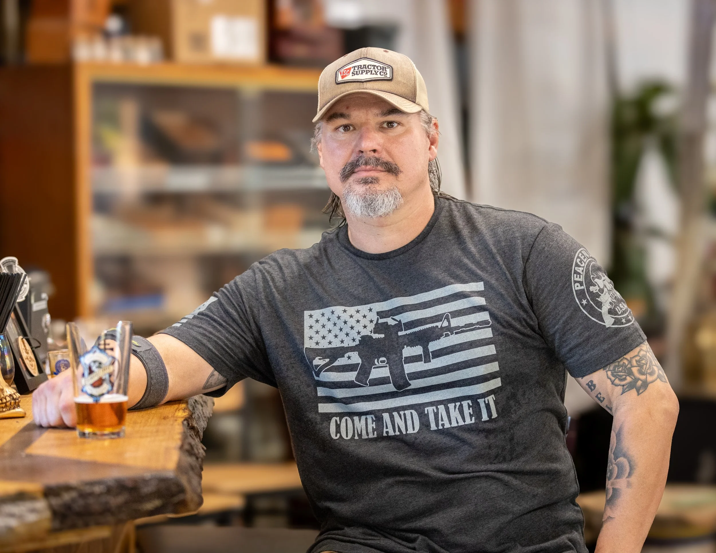 A man with a beard and mustache wearing a beige baseball cap and a black T-shirt with a gun graphic and the phrase "Come and Take It" standing at a bar with a shot of whiskey in front of him. Shelves with various items are blurred in the background.