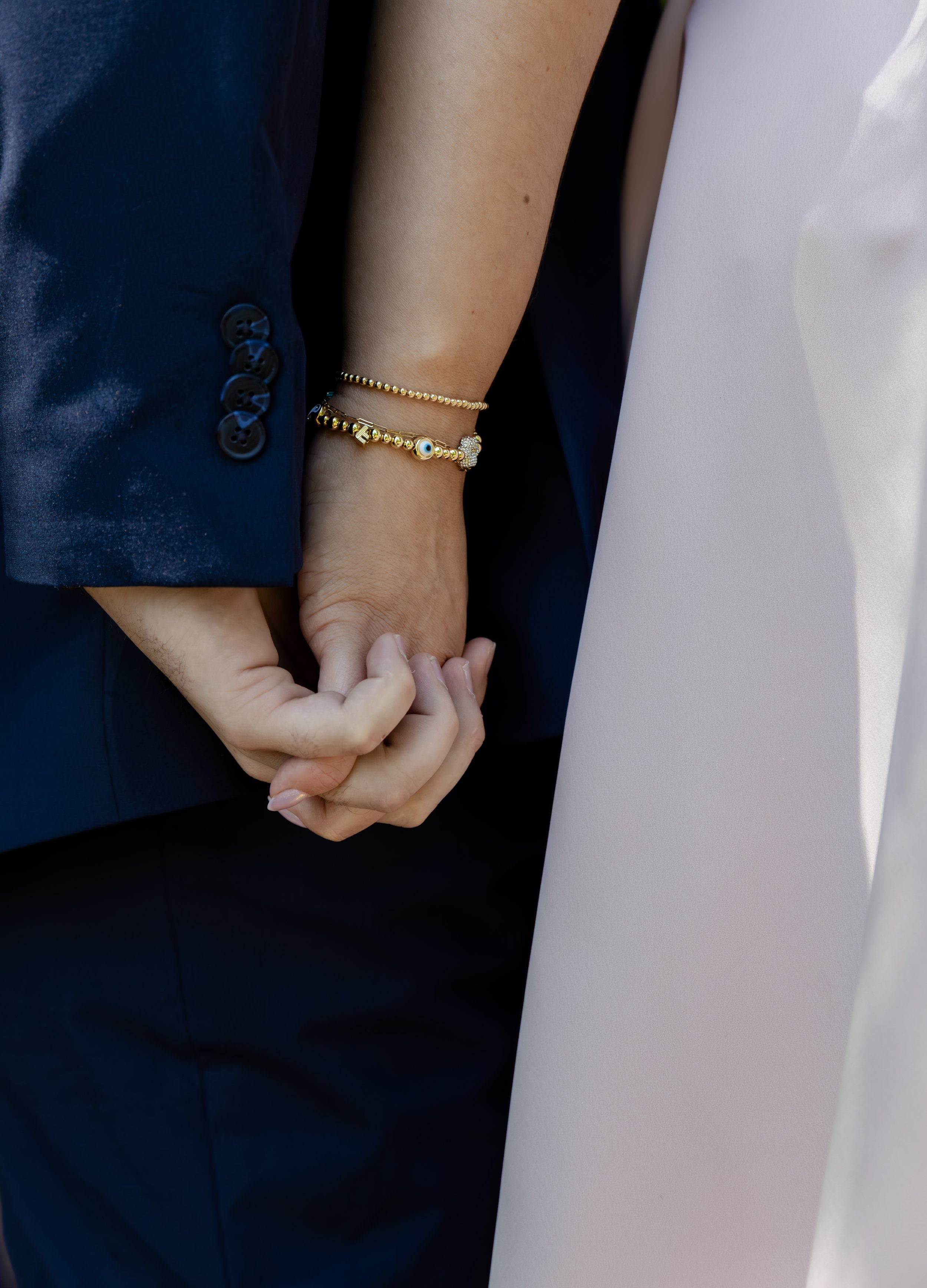 A close-up of two people holding hands. One person is wearing a dark blue suit and multiple bracelets, and the other is wearing light-colored clothing.