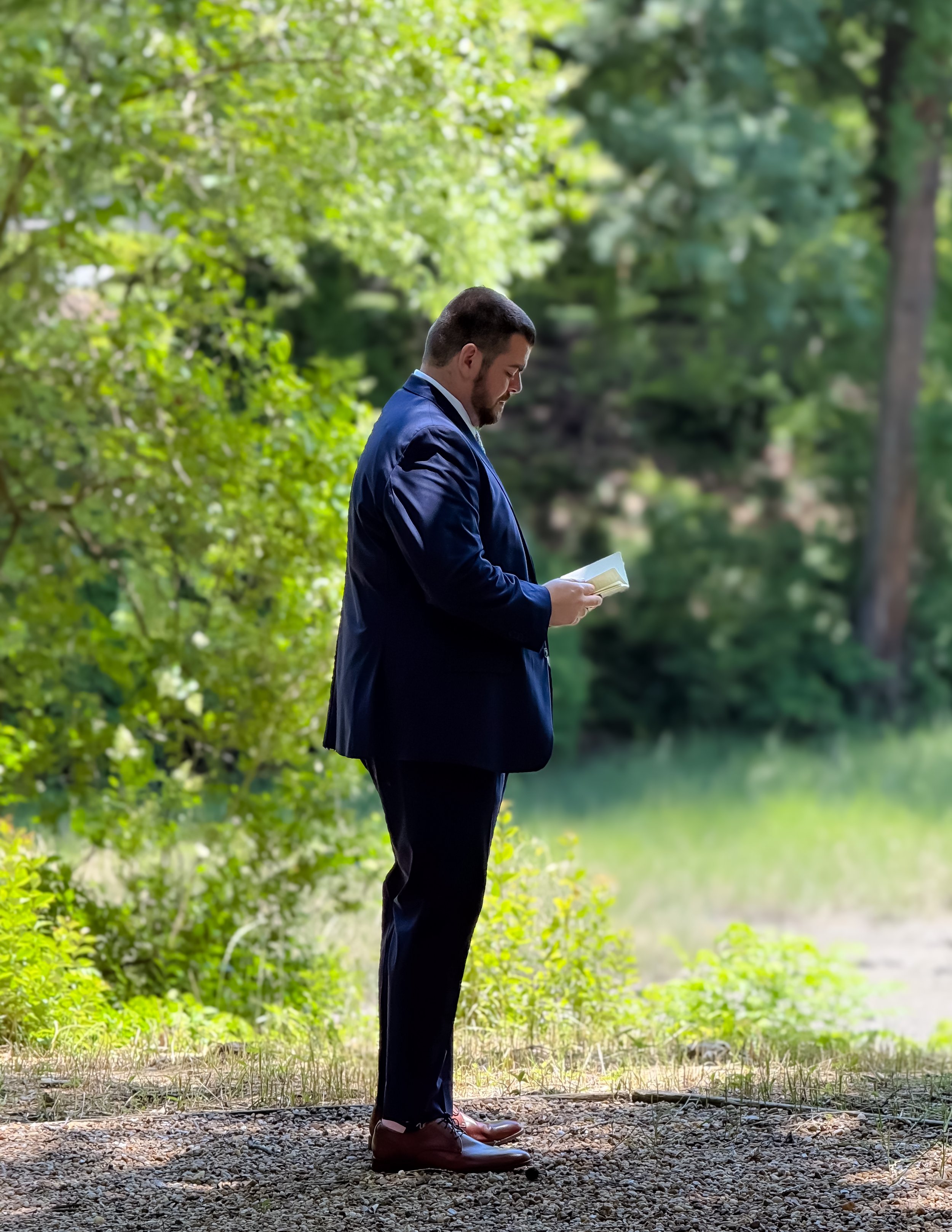 A man in a blue suit reading a booklet outdoors in a wooded area.