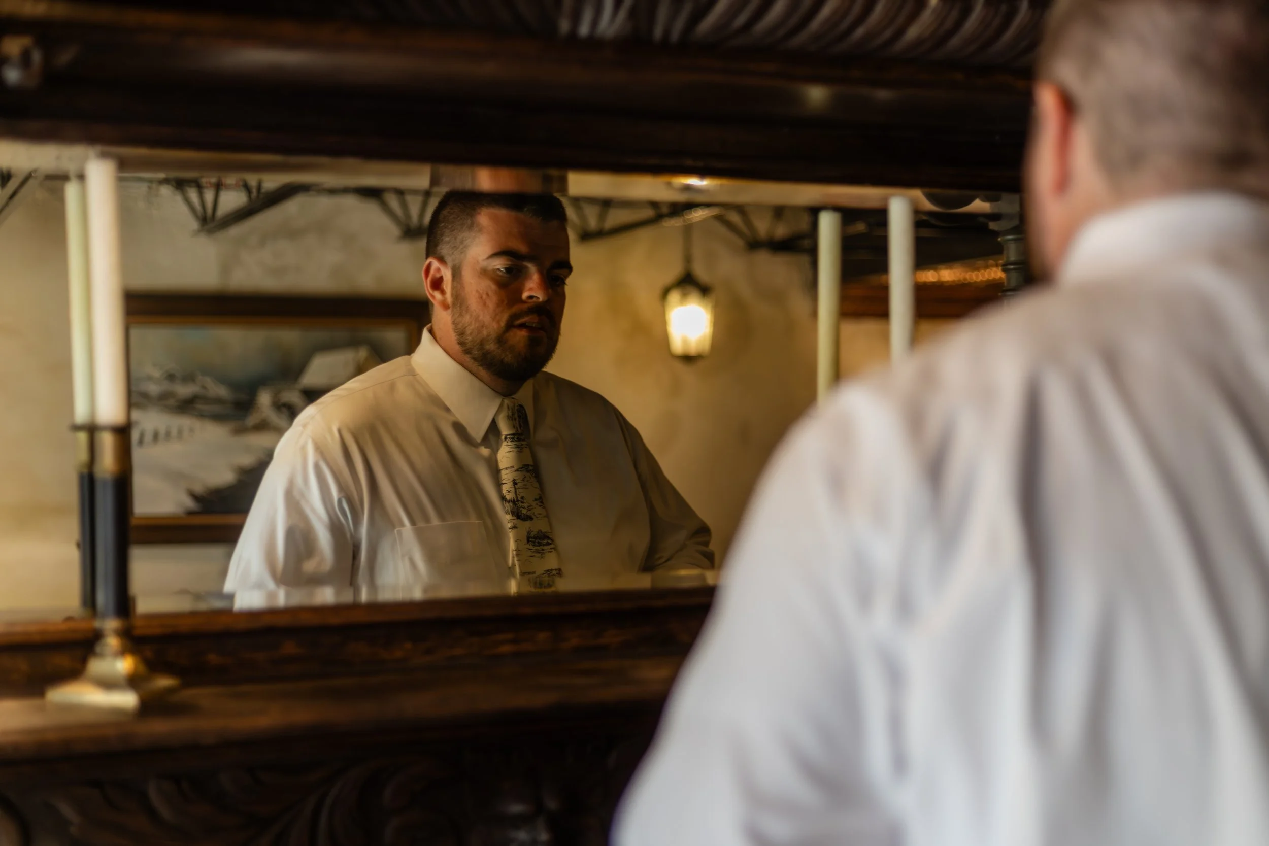 Man with short dark hair and beard looking into a mirror, wearing a cream-colored shirt and patterned tie, in a warmly lit room.