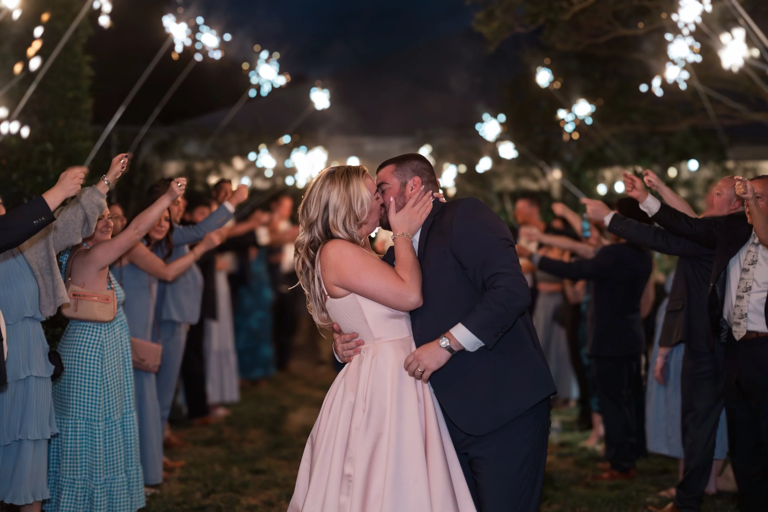 Couple sharing a kiss during their wedding reception under string lights, with guests holding sparklers on either side.