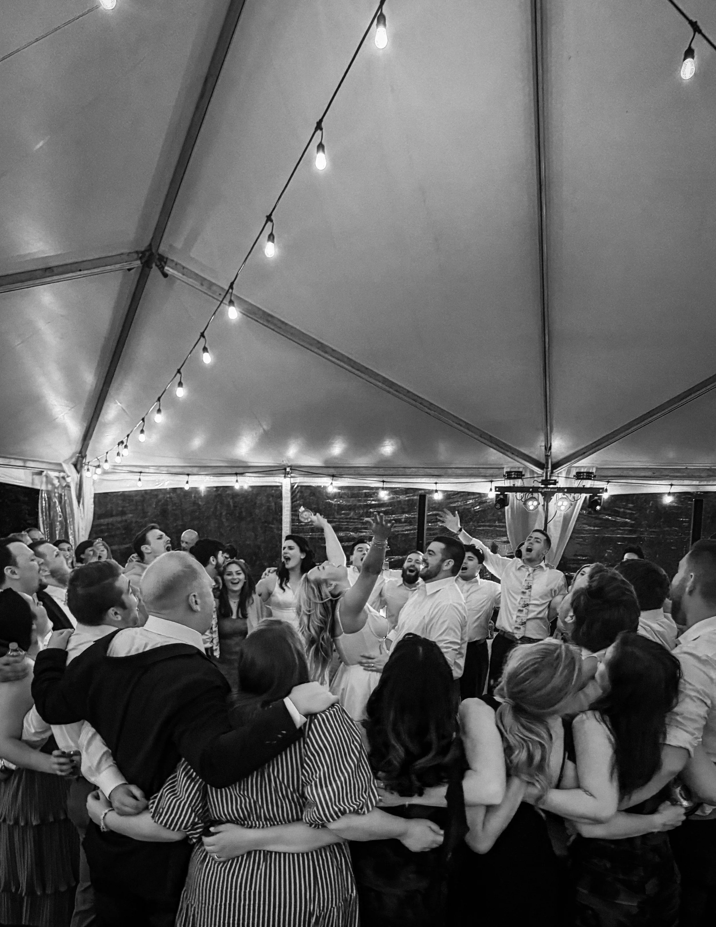 A group of people celebrating at a wedding reception inside a tent with string lights, dancing and embracing each other.
