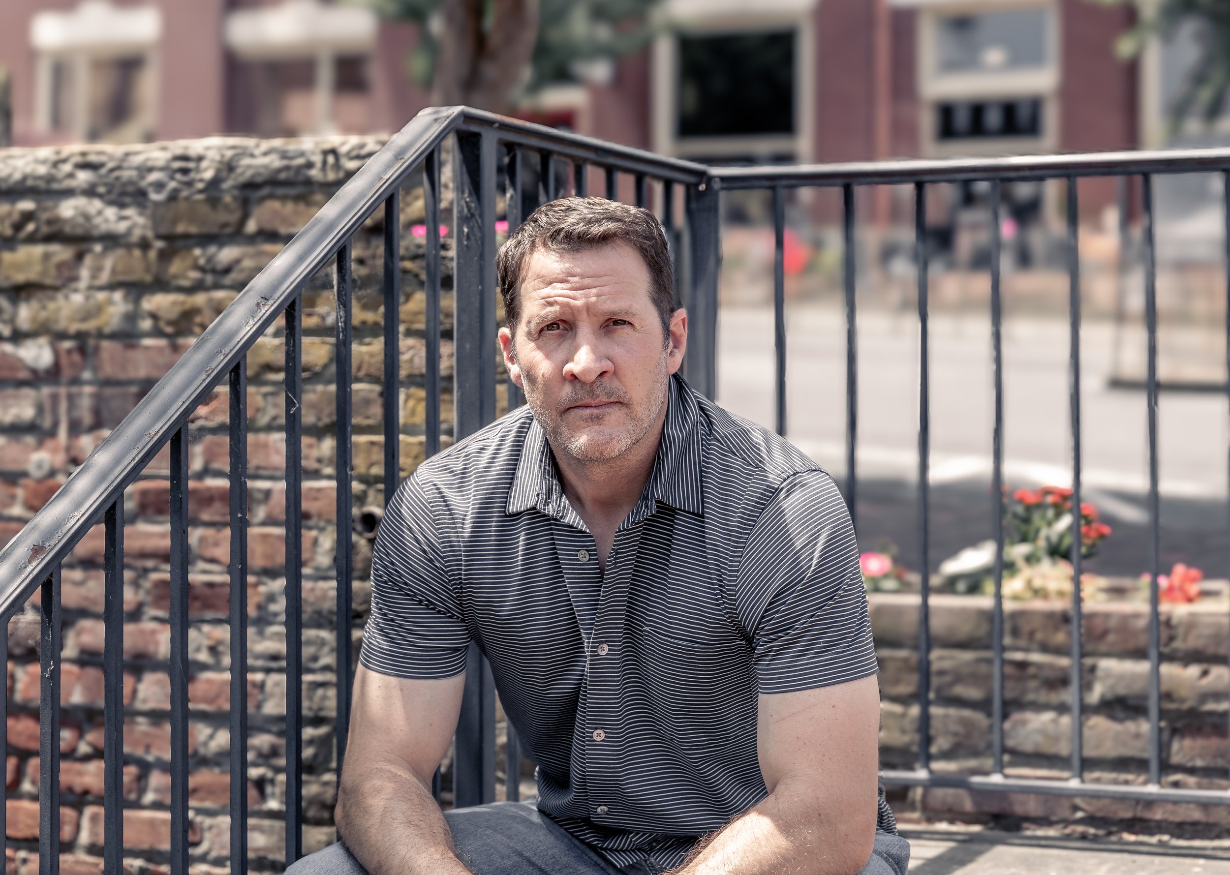 A man with short dark hair, a light beard, and a striped polo shirt sitting outdoors near a brick wall and metal railing, with a blurred background of a city street and buildings.
