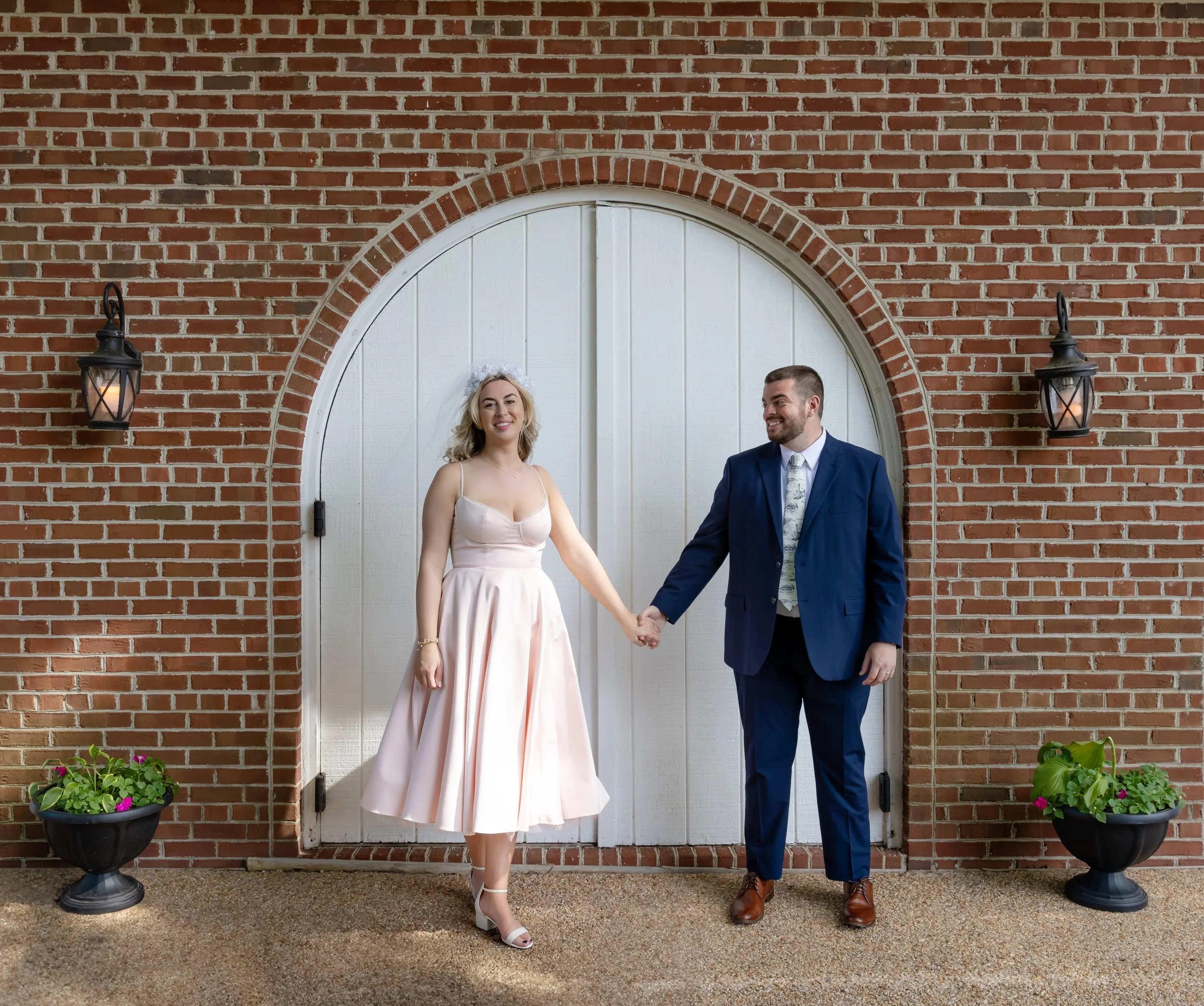 A bride and groom holding hands in front of a white arched door framed by a brick wall, with two black lantern-style lights and potted plants on either side.