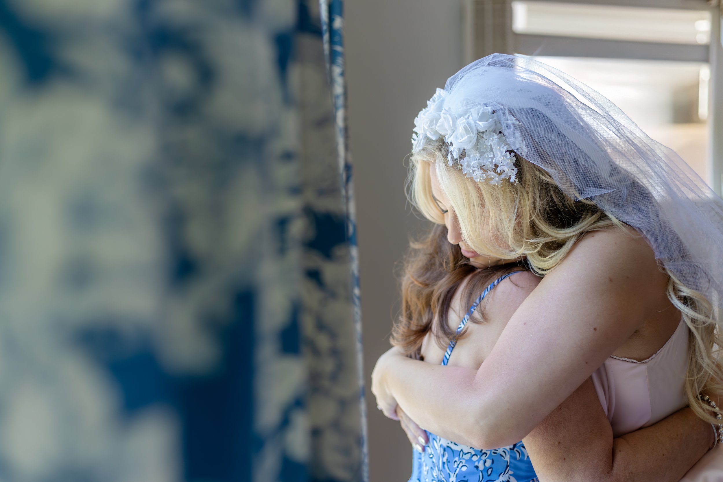 Bride wearing a veil and floral headpiece hugging a woman in a blue dress.