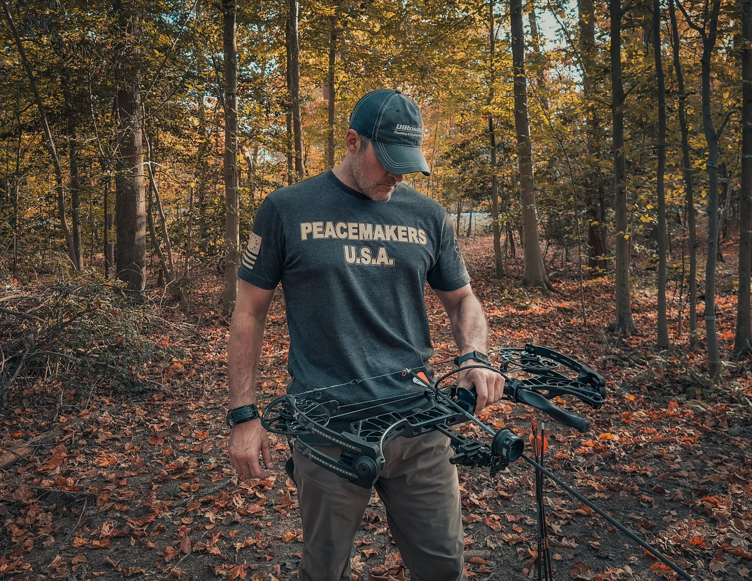 A man stands in a forested area during autumn, holding a black compound bow, wearing a dark gray t-shirt with "PEACEMAKERS U.S.A." printed on it, a baseball cap, and beige pants.