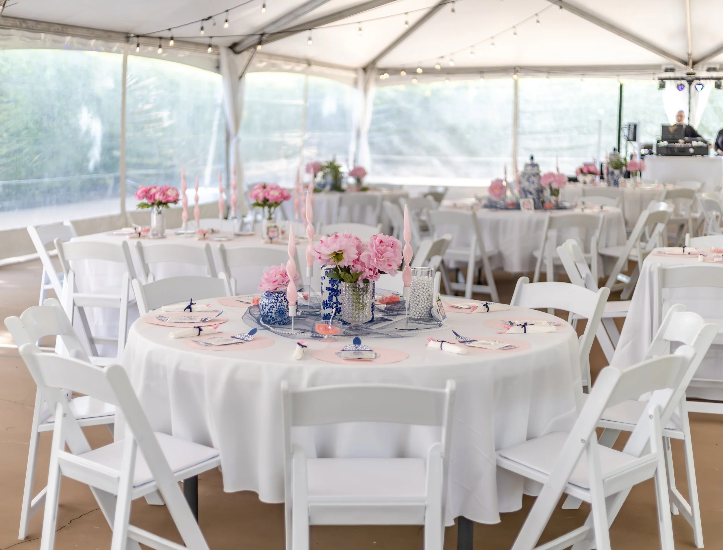 Tea party or wedding reception setup under a tent with white tables, pink floral centerpieces, pink candles, and white chairs.