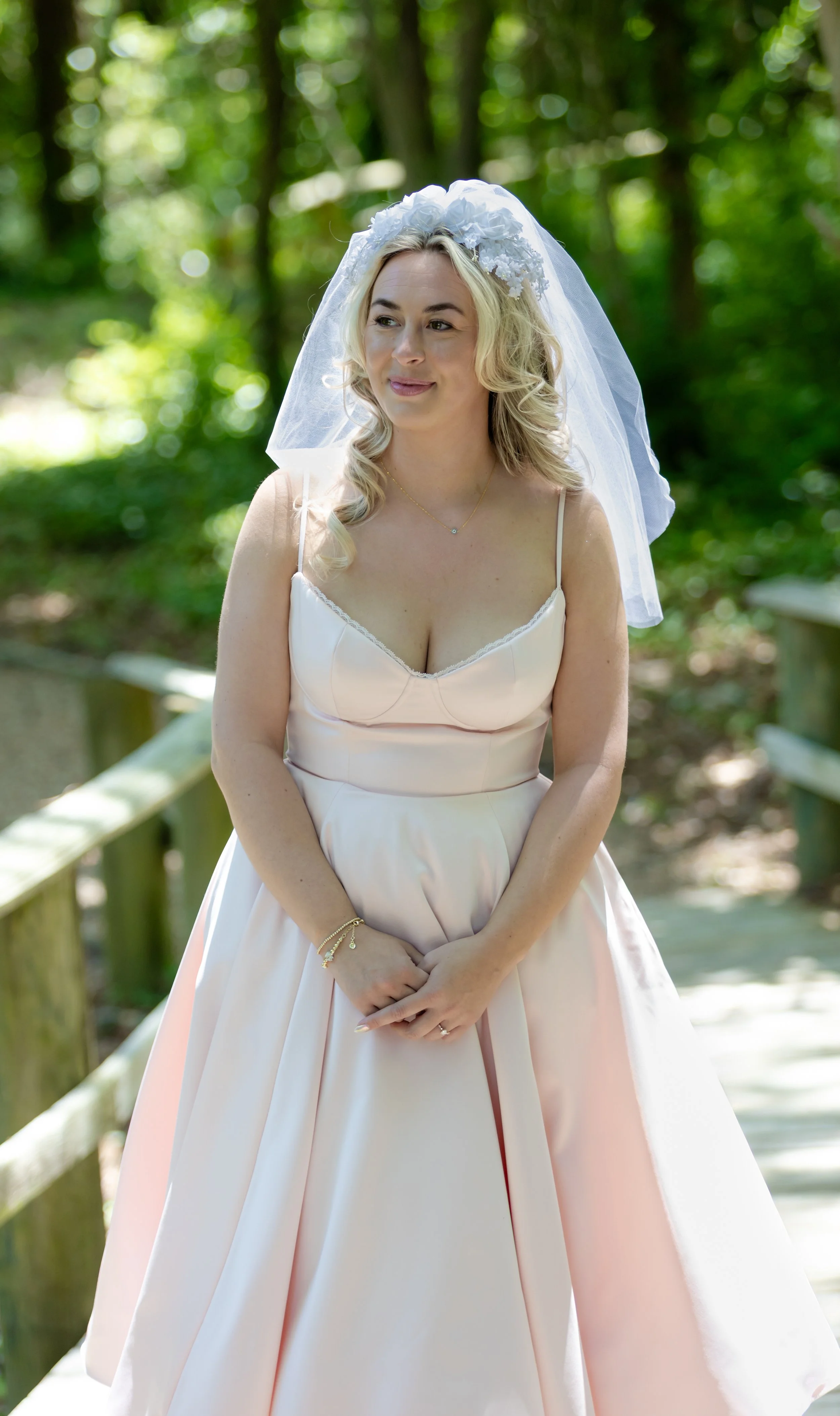 A woman dressed in a light pink dress with spaghetti straps, wearing a bridal veil, standing on a wooden pathway surrounded by green trees.