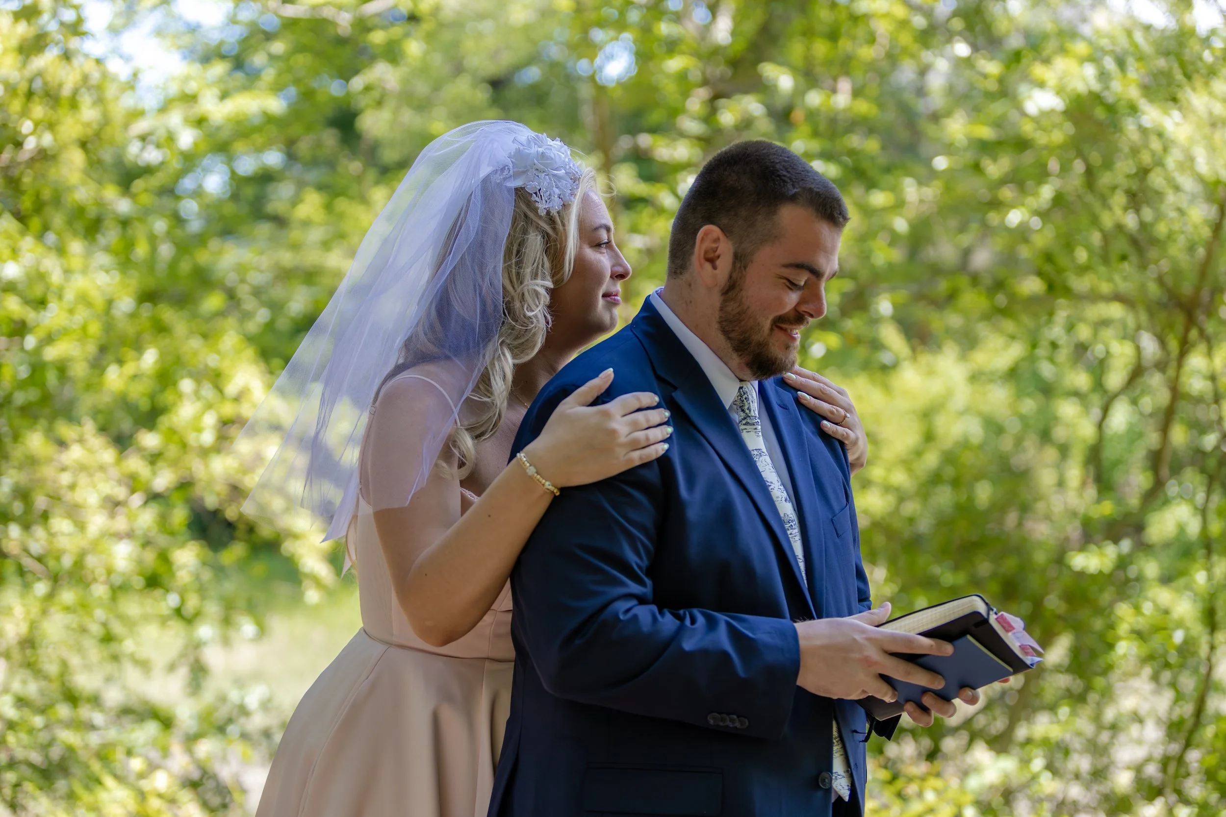 A bride and groom standing outdoors during a wedding ceremony, with the bride hugging the groom from behind as he reads from a book.