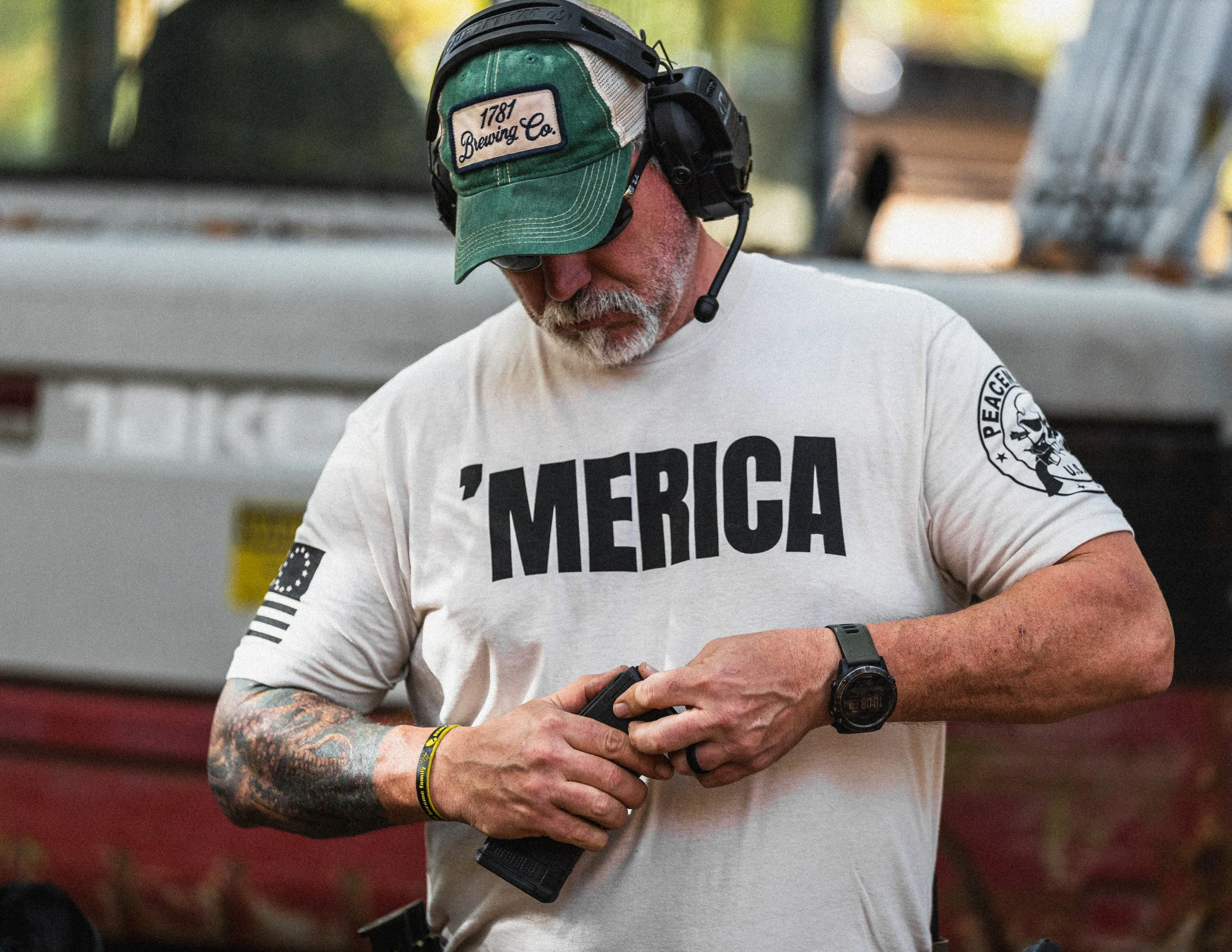 A man wearing a green baseball cap and a white t-shirt with the word 'MERICA' standing outdoors, looking at his phone with a headset on and a watch on his wrist, tattoos on his arms, and a patch with a skull and peace sign on his sleeve.