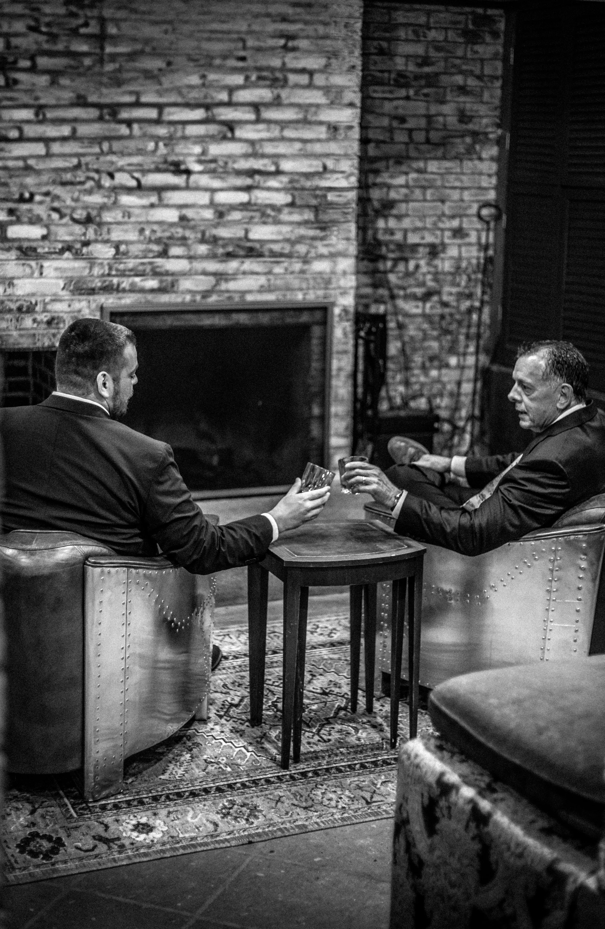 Two men in suits sitting on armchairs, drinking from glasses, engaged in conversation in a cozy room with exposed brick wall and fireplace.