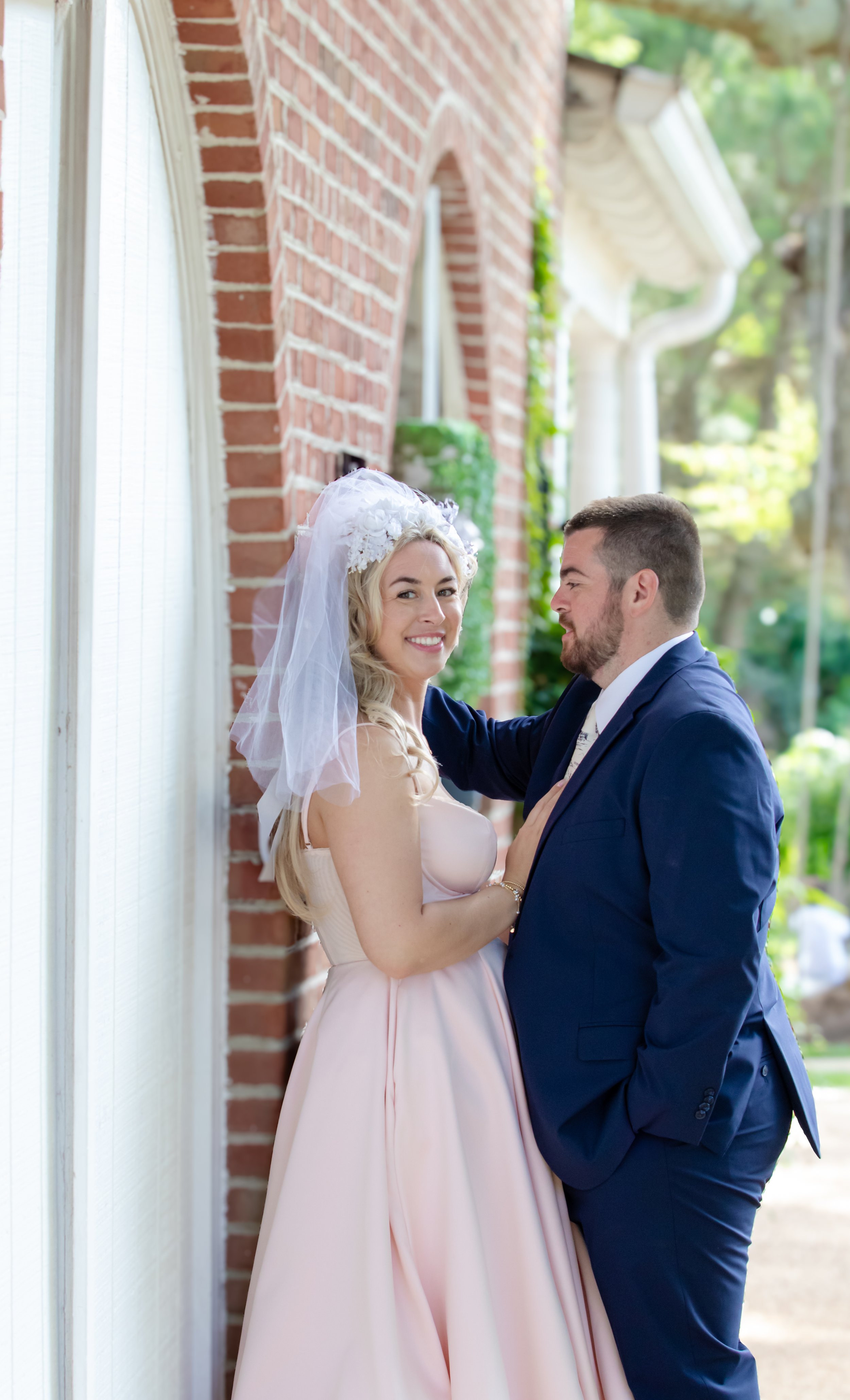 A bride and groom sharing a moment outside near a brick building, dressed in wedding attire, with the bride in a pink gown and veil, and the groom in a navy suit.