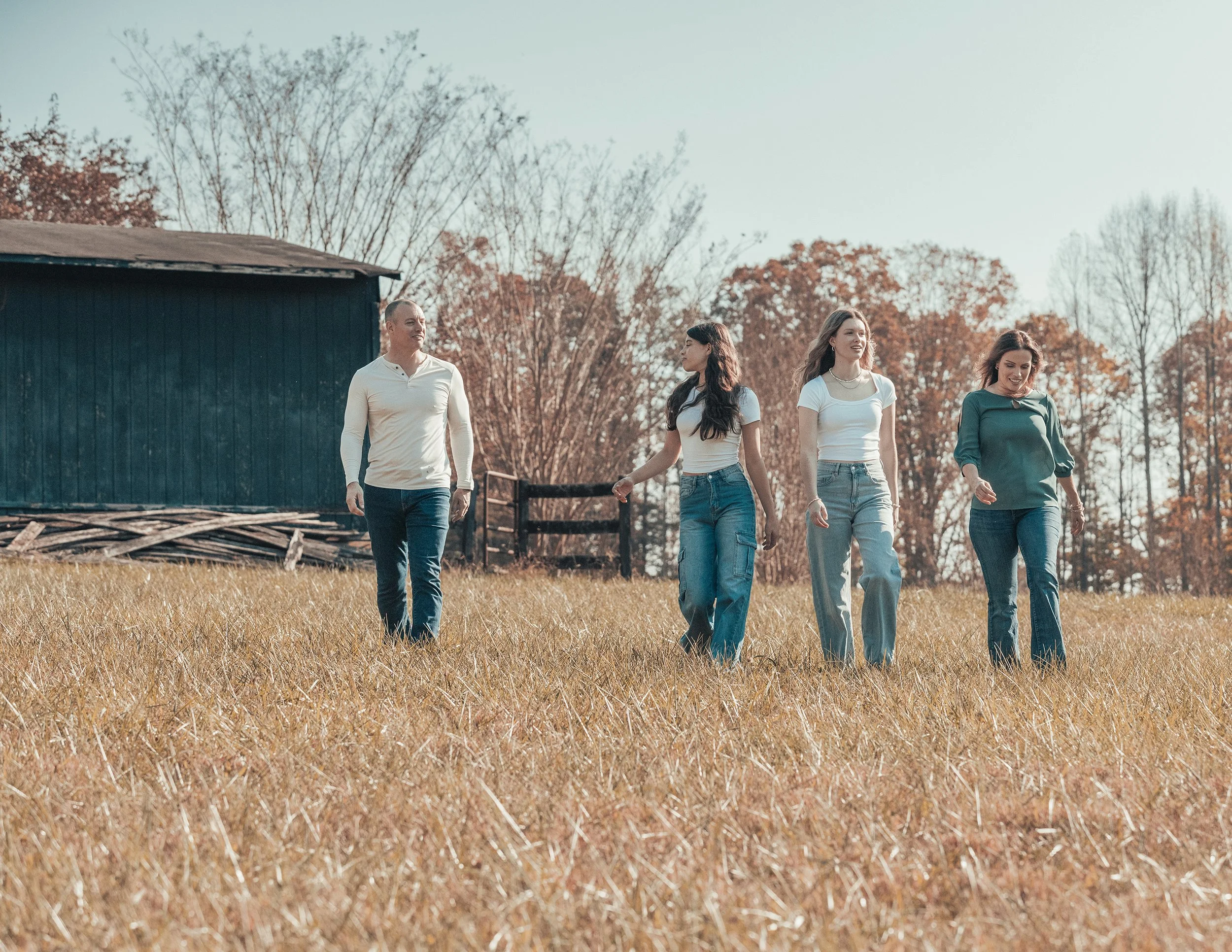 Four people walking through a grassy field in autumn with trees and a blue barn in the background.