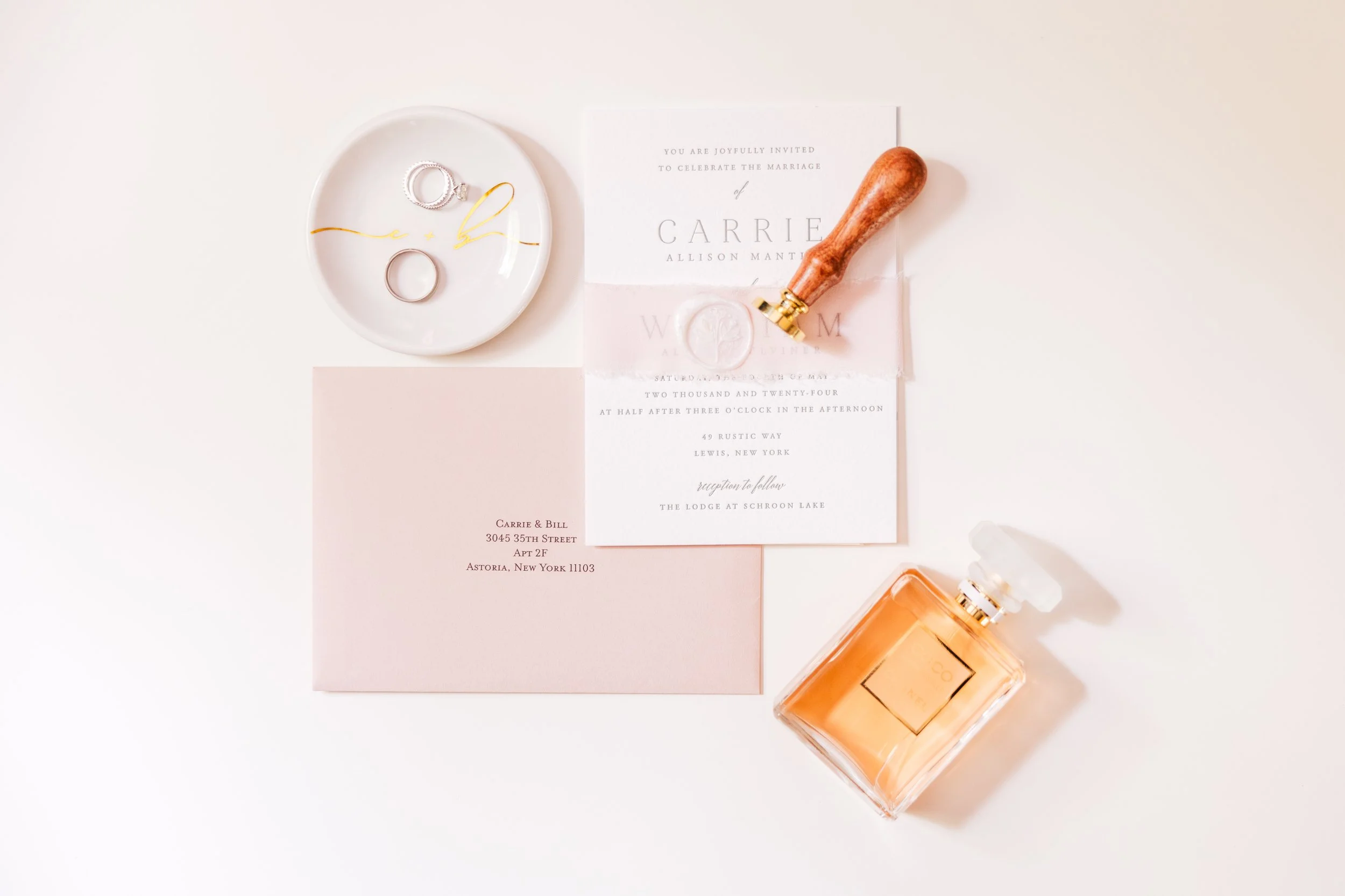 Invitation to a wedding, rings on a white dish, a pink envelope, a sealing wax stamp, a wooden stamp, and a perfume bottle on a white background.